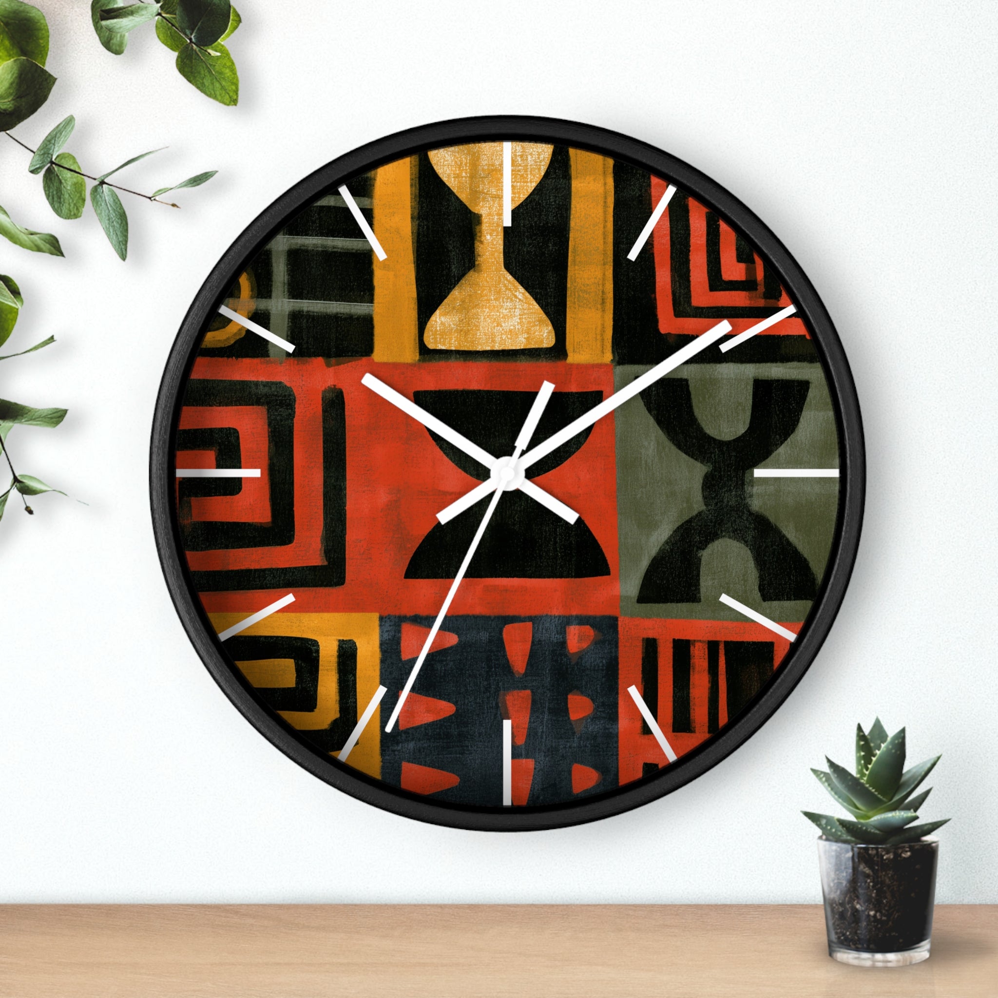 Black Afrocentric abstract geometric round wall clock with white time markings and white clock hands, featuring African-inspired symbols, spirals, arches, stripes, and graphic patterns in red, black, mustard gold, charcoal, and earthy gray, displayed against a white background with plant accessories.
