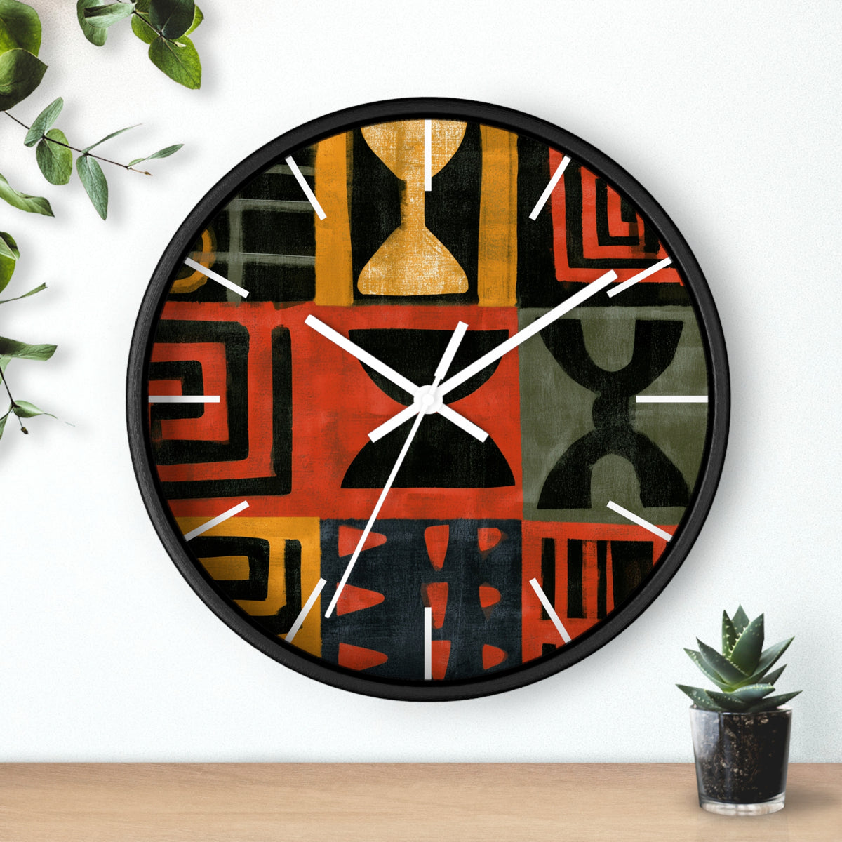 Black Afrocentric abstract geometric round wall clock with white time markings and white clock hands, featuring African-inspired symbols, spirals, arches, stripes, and graphic patterns in red, black, mustard gold, charcoal, and earthy gray, displayed against a white background with plant accessories.