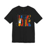 A black short-sleeve crewneck t-shirt featuring a large graphic on the chest spelling 'JUSTICE' in stylized block letters filled with red, yellow, blue, and white patterns, shown against a plain white background.