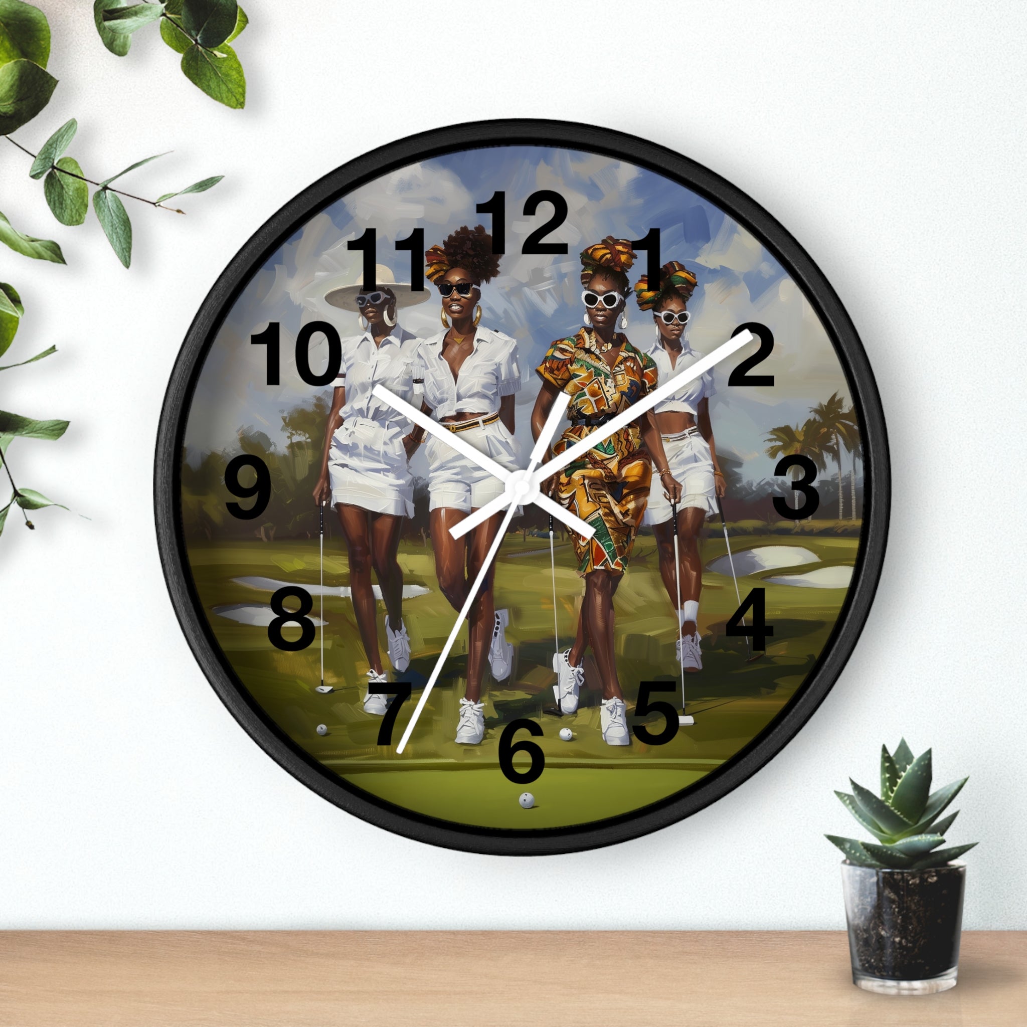 A round wall clock with a black frame and white hands, featuring a printed illustration of three women golfing on a green course. The clock is mounted on a white wall above a light wood surface, with decorative green leaves and a small potted succulent nearby.