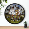 A round wall clock with a black frame and white hands, featuring a printed illustration of three women golfing on a green course. The clock is mounted on a white wall above a light wood surface, with decorative green leaves and a small potted succulent nearby.