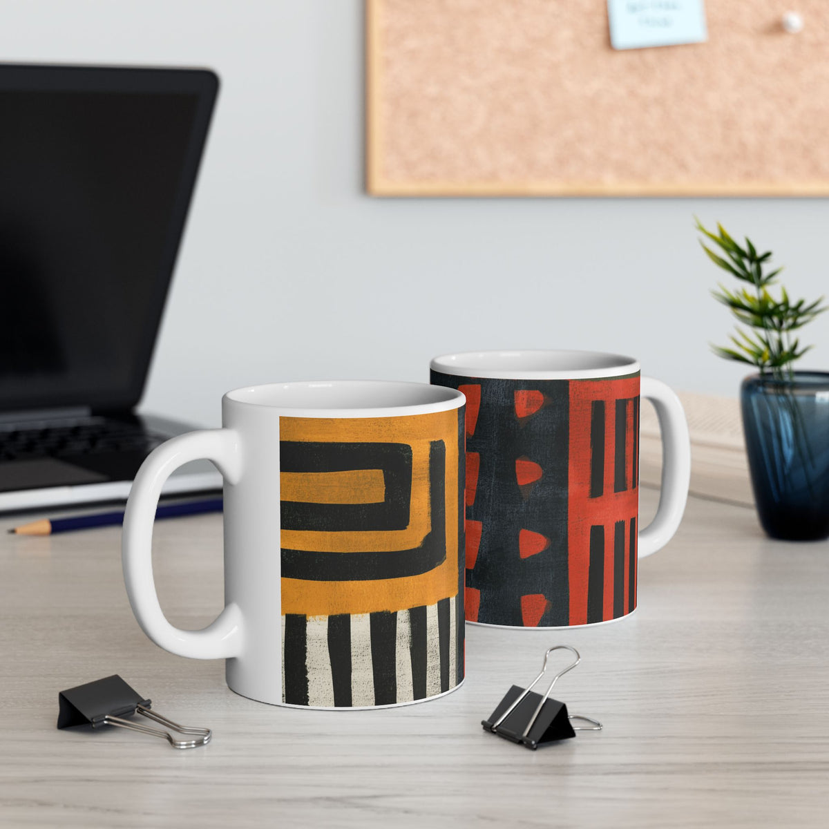 Left and right handle views of 11 oz. red, black, gold and cream abstract art coffee mug featuring bold geometric circles, stripes, organic shapes and a Bauhaus-inspired modern design, displayed on top of an office desk with a laptop computer in the background.
