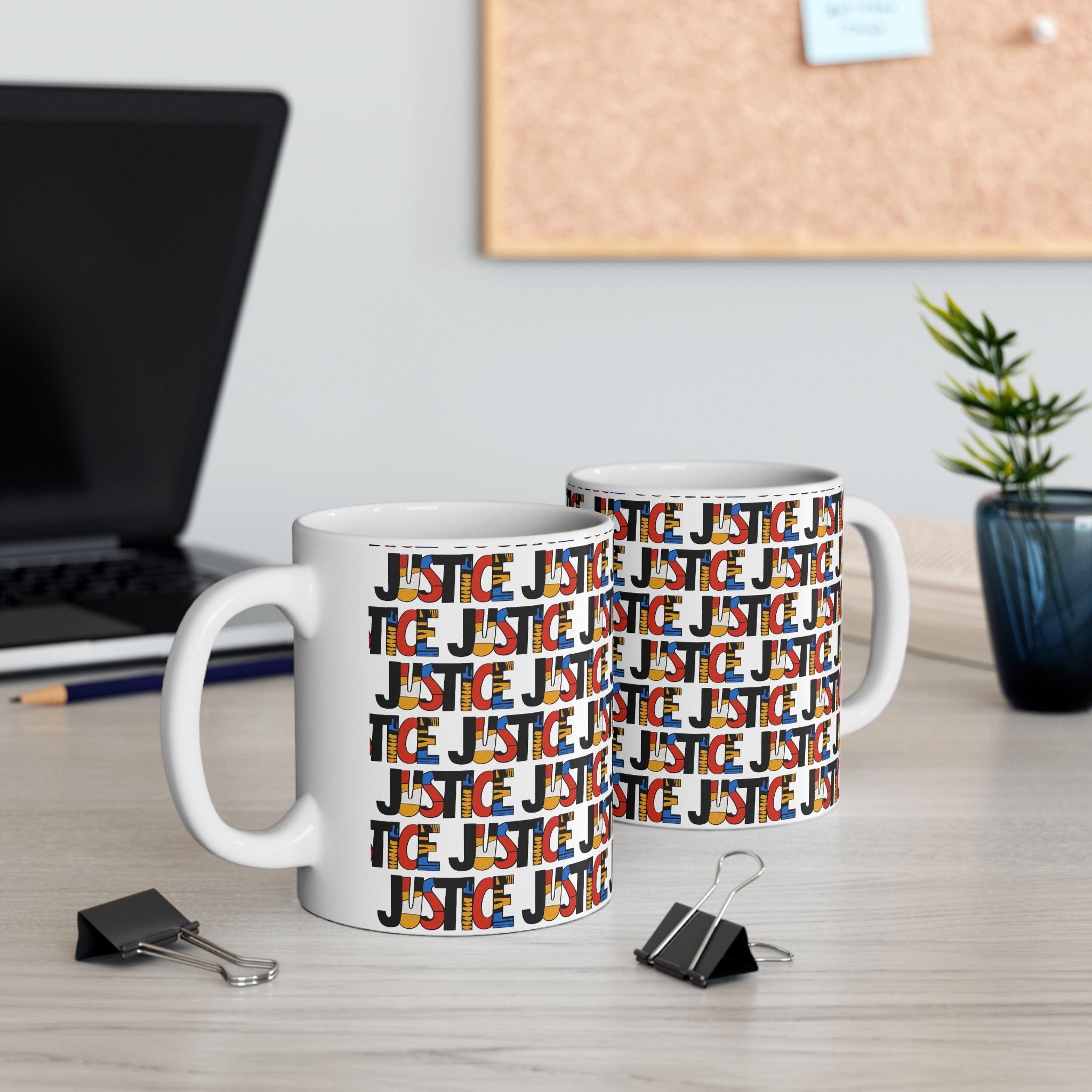 Two white ceramic mugs with a repeating 'JUSTICE' graphic pattern in red, orange, yellow, and black, displayed on a light wood desk in an office setting with a laptop, binder clips, and a plant in the background.
