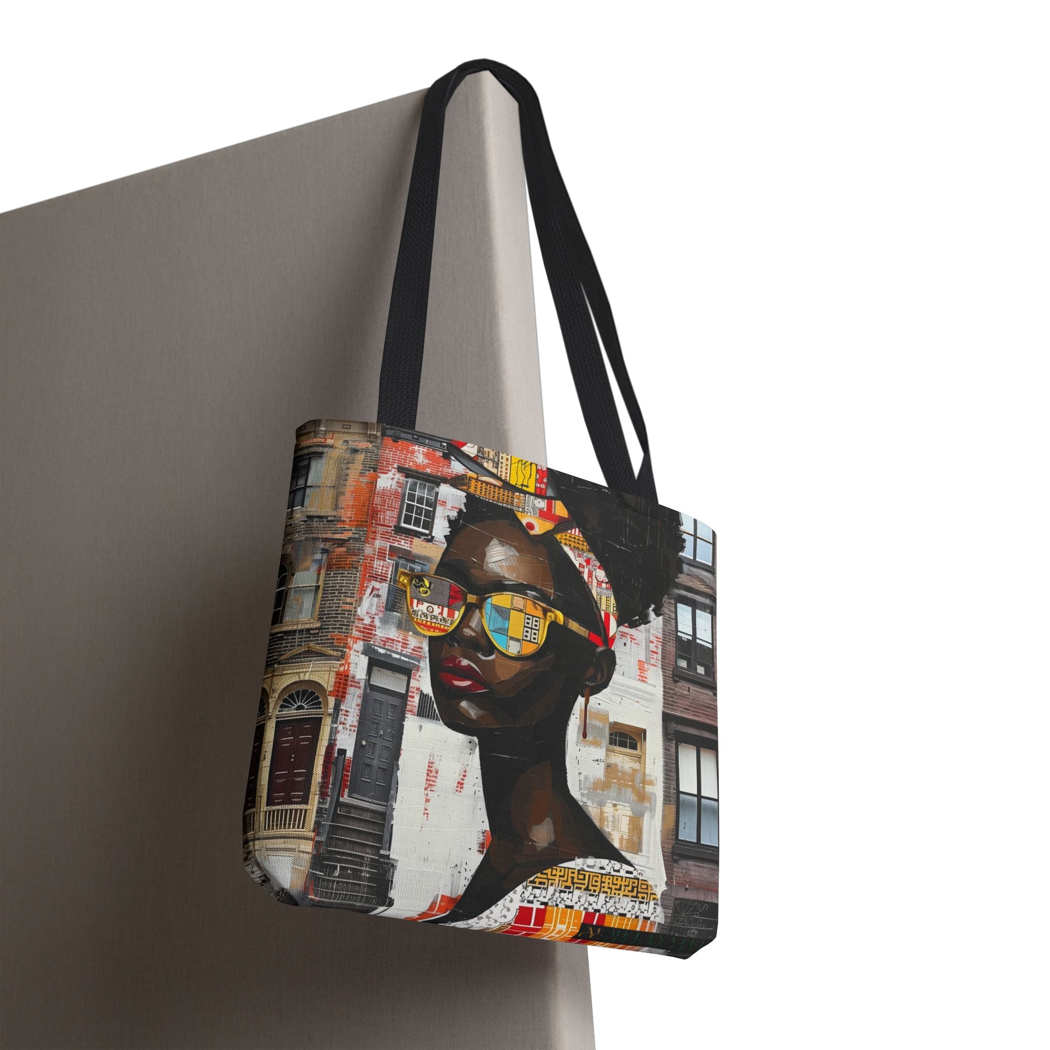 A tote bag with black shoulder straps, featuring a colorful artistic print of a Black woman wearing yellow sunglasses against a city street background, shown hanging against a neutral gray wall.