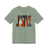 A sage green crewneck t-shirt featuring a large graphic print on the front spelling 'JUSTICE' in bold, multi-colored geometric patterns, shown against a plain white background.