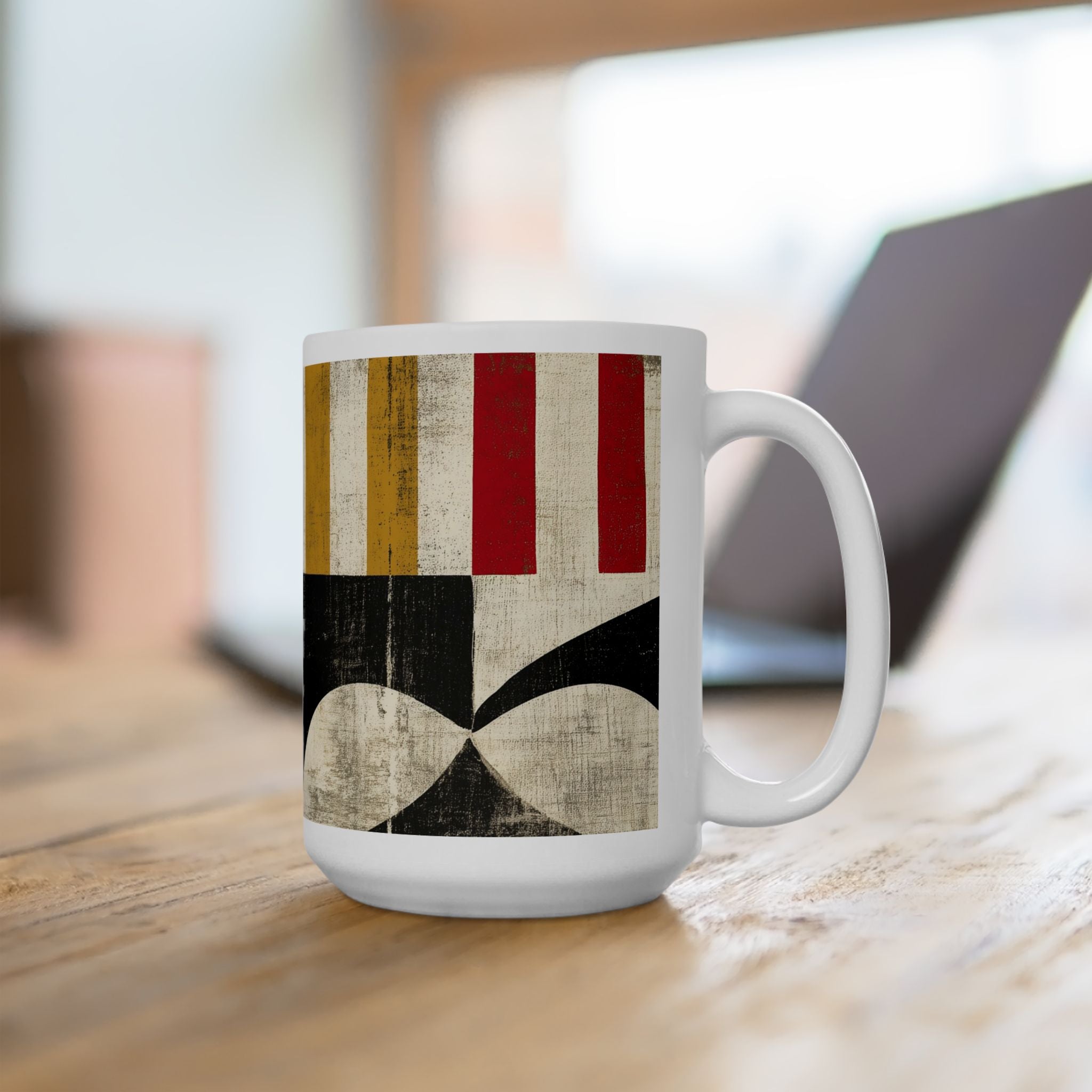 A 15 oz white ceramic mug with a vintage-style geometric pattern featuring red, gold, black, and beige stripes and shapes, sitting on a wooden table with a blurred laptop in the background.