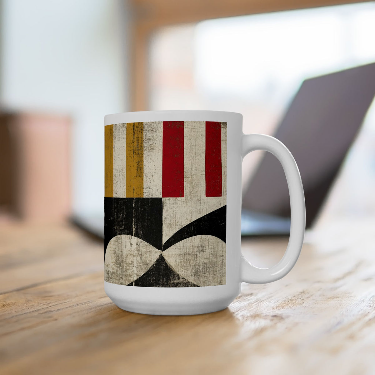 A 15 oz white ceramic mug with a vintage-style geometric pattern featuring red, gold, black, and beige stripes and shapes, sitting on a wooden table with a blurred laptop in the background.