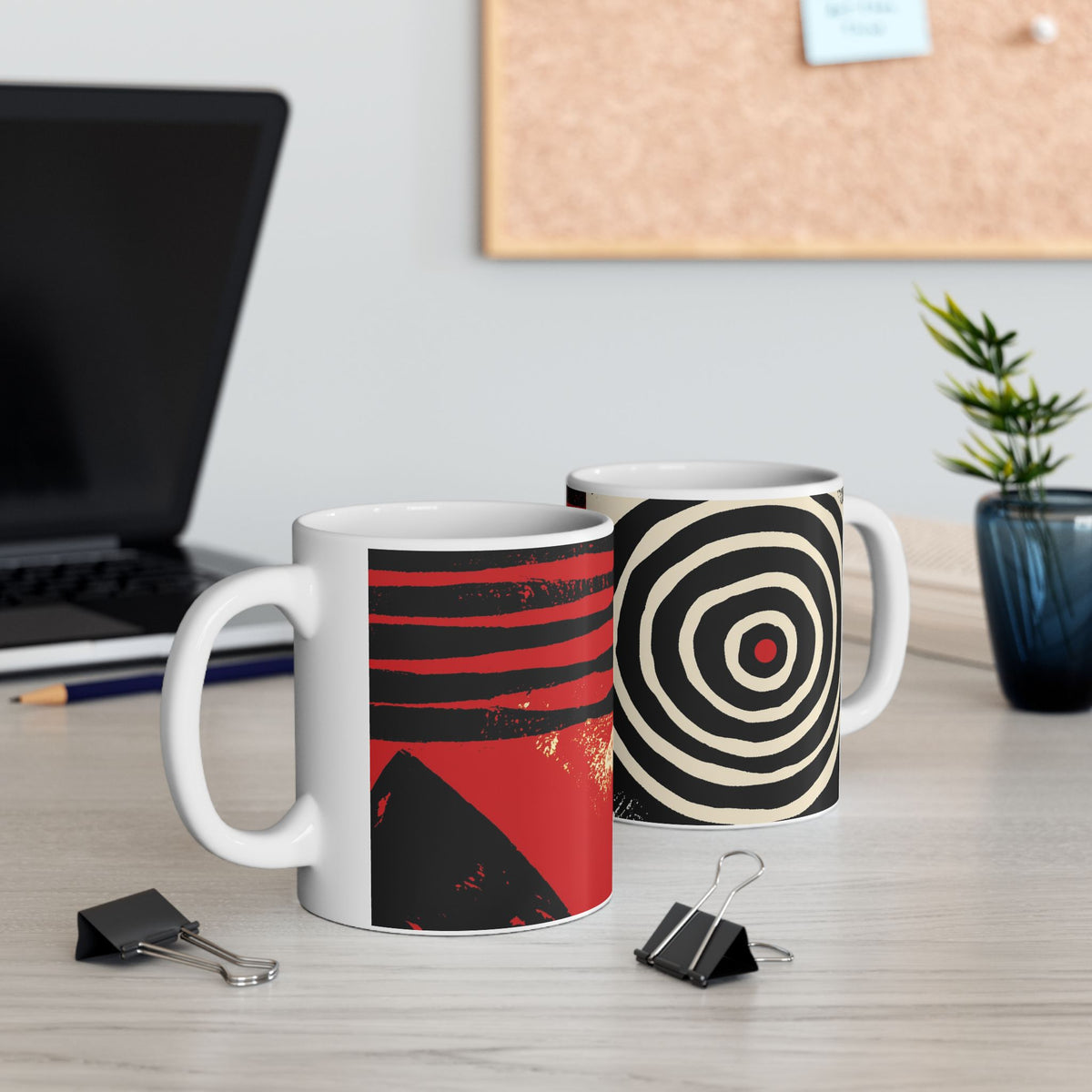Left and right handle views of 11 oz. red, black and cream abstract art coffee mug featuring bold geometric circles, stripes, organic shapes and a Bauhaus-inspired modern design, displayed on top of an office desk with a laptop computer in the background.