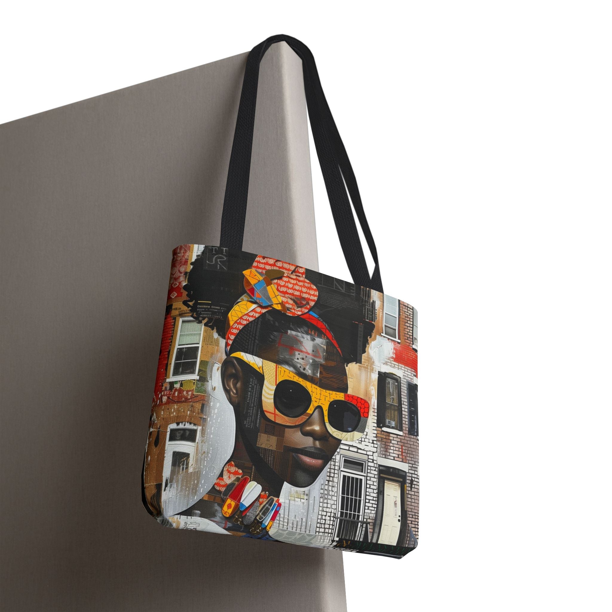 A tote bag with black shoulder straps, featuring a detailed graphic print of a black woman wearing yellow sunglasses and a headwrap, set against a collage of city buildings. The bag is shown hanging against a neutral gray background.