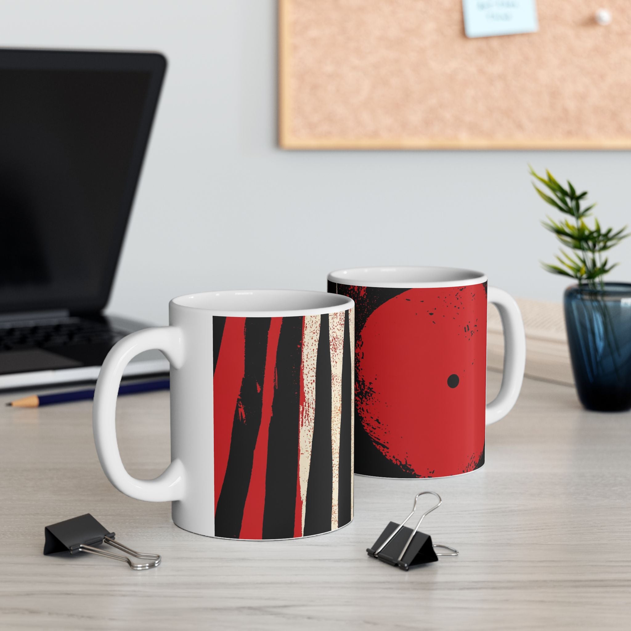 Left and right handle views of 11 oz. red, black and cream abstract art coffee mug featuring bold geometric circles, stripes, organic shapes and a Bauhaus-inspired modern design, displayed on top of an office desk with a laptop computer in the background.
