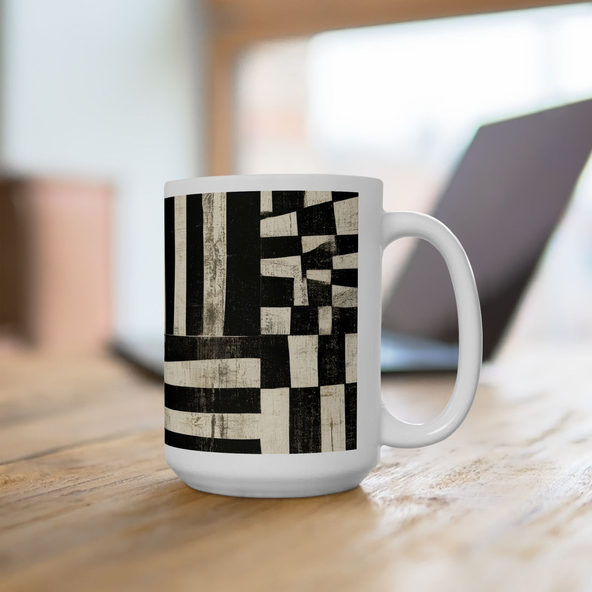 A white 15 oz. ceramic mug with a black and beige geometric block pattern, sitting on a wooden table with a blurred laptop in the background.