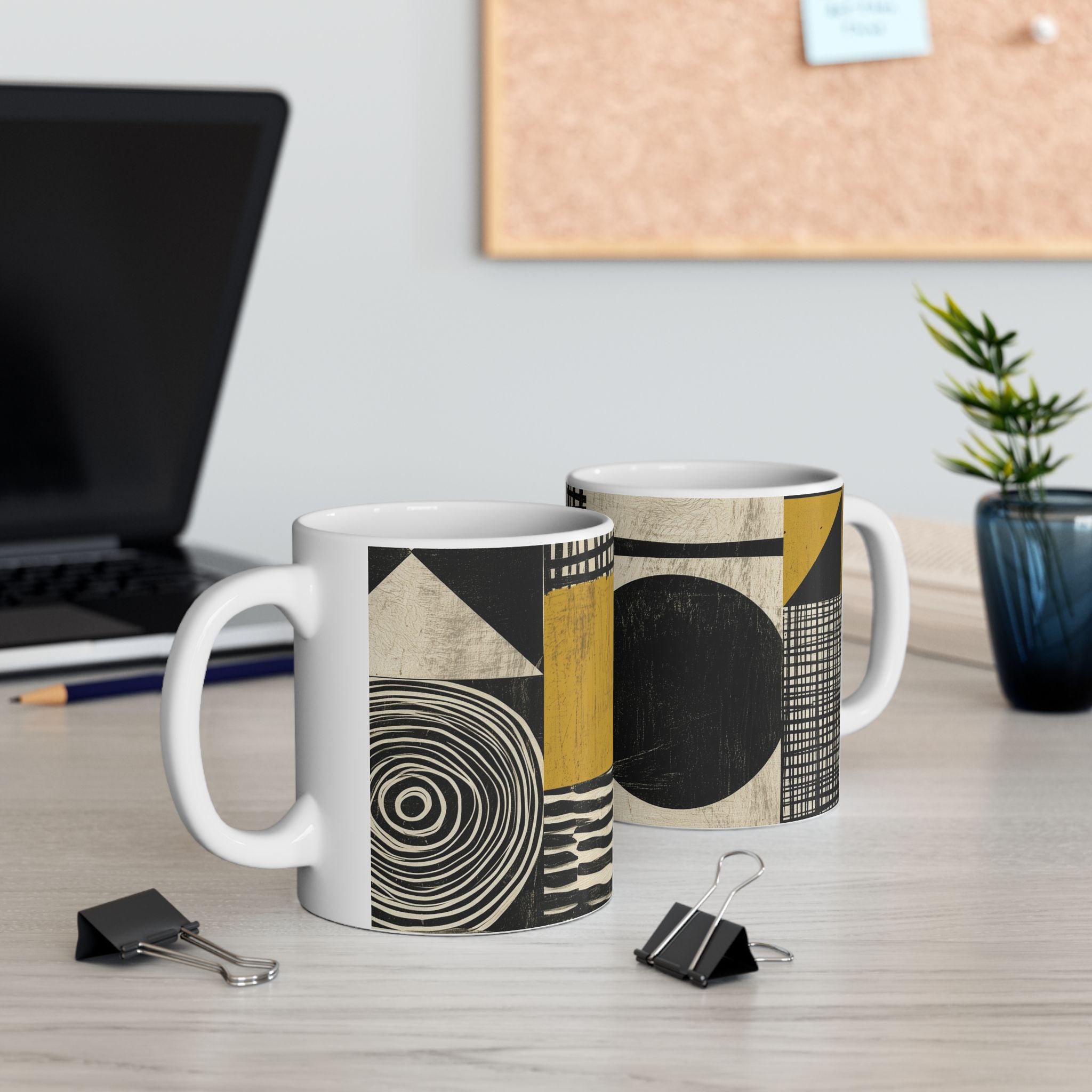 Two white ceramic mugs with a modern abstract geometric pattern in black, beige, and mustard yellow, displayed on a light wood desk with office props like a laptop, binder clips, and a plant.