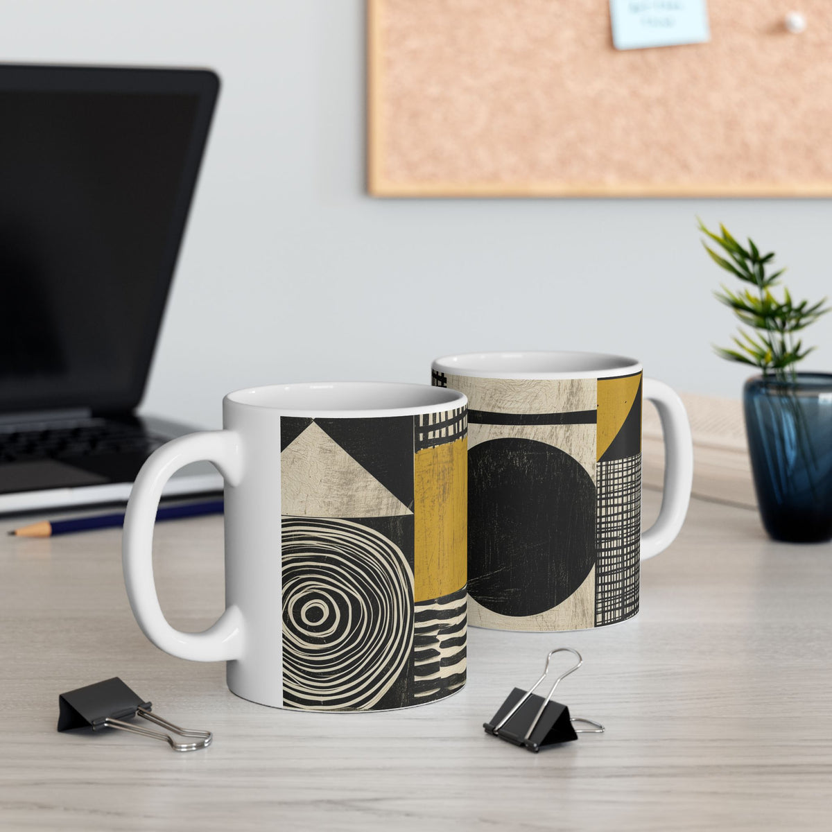 Two white ceramic mugs with a modern abstract geometric pattern in black, beige, and mustard yellow, displayed on a light wood desk with office props like a laptop, binder clips, and a plant.