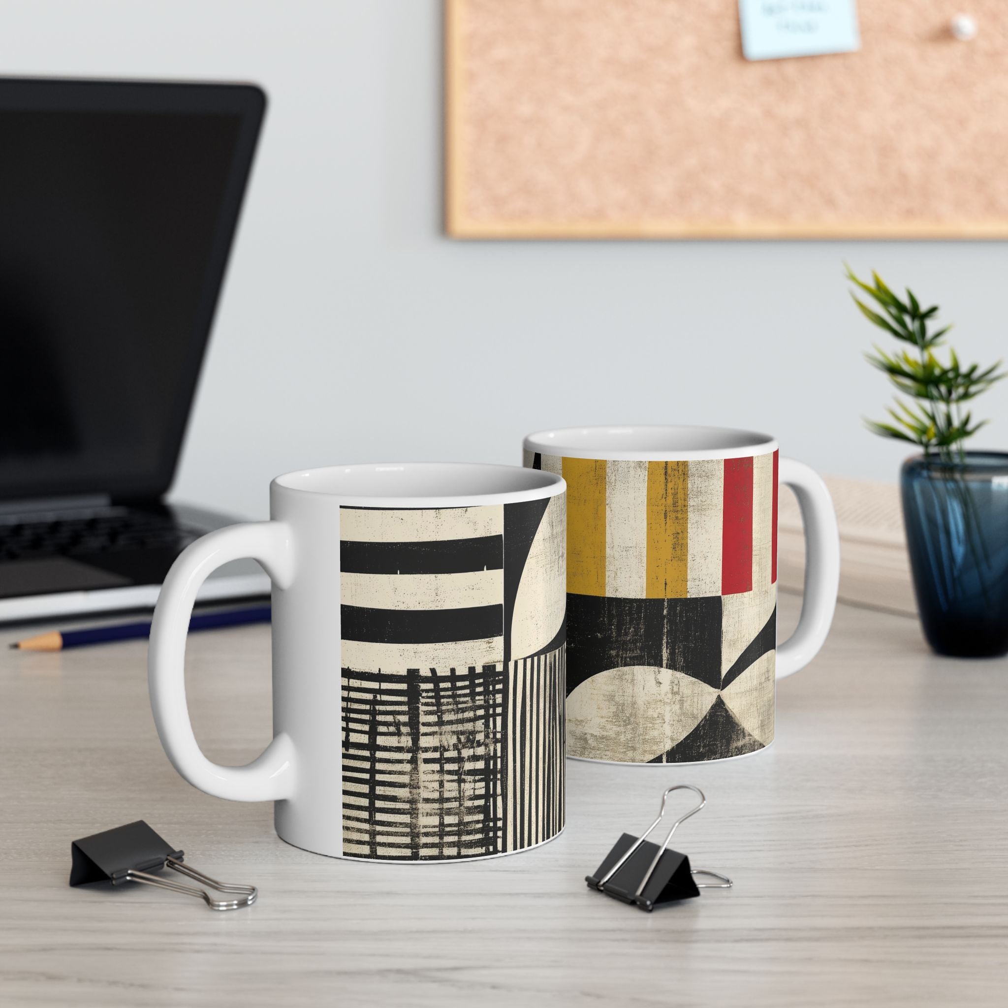 Two ceramic mugs with abstract geometric patterns displayed on a light wood desk in an office setting, with a laptop, binder clips, and a small plant in the background.