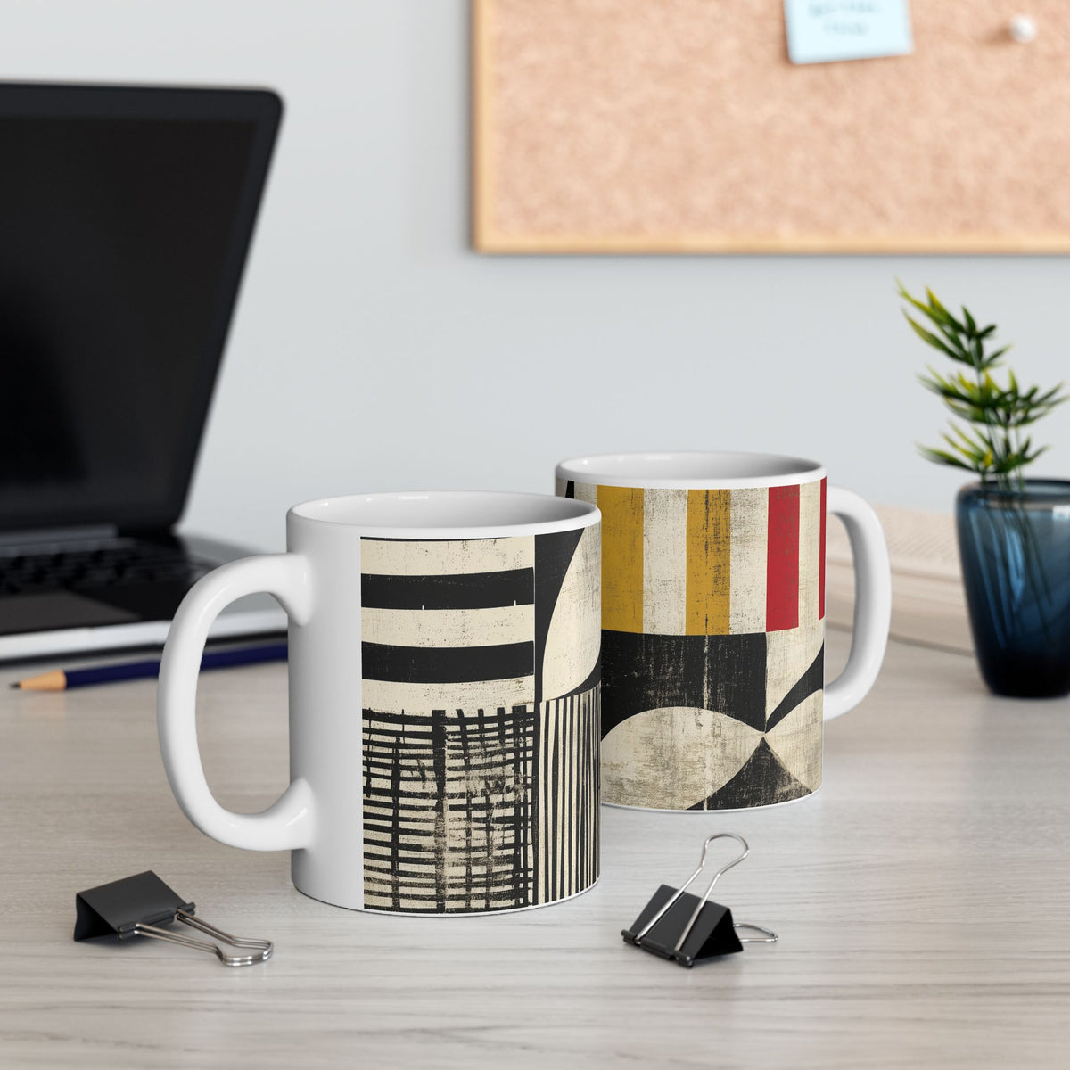 Two ceramic mugs with abstract geometric patterns displayed on a light wood desk in an office setting, with a laptop, binder clips, and a small plant in the background.