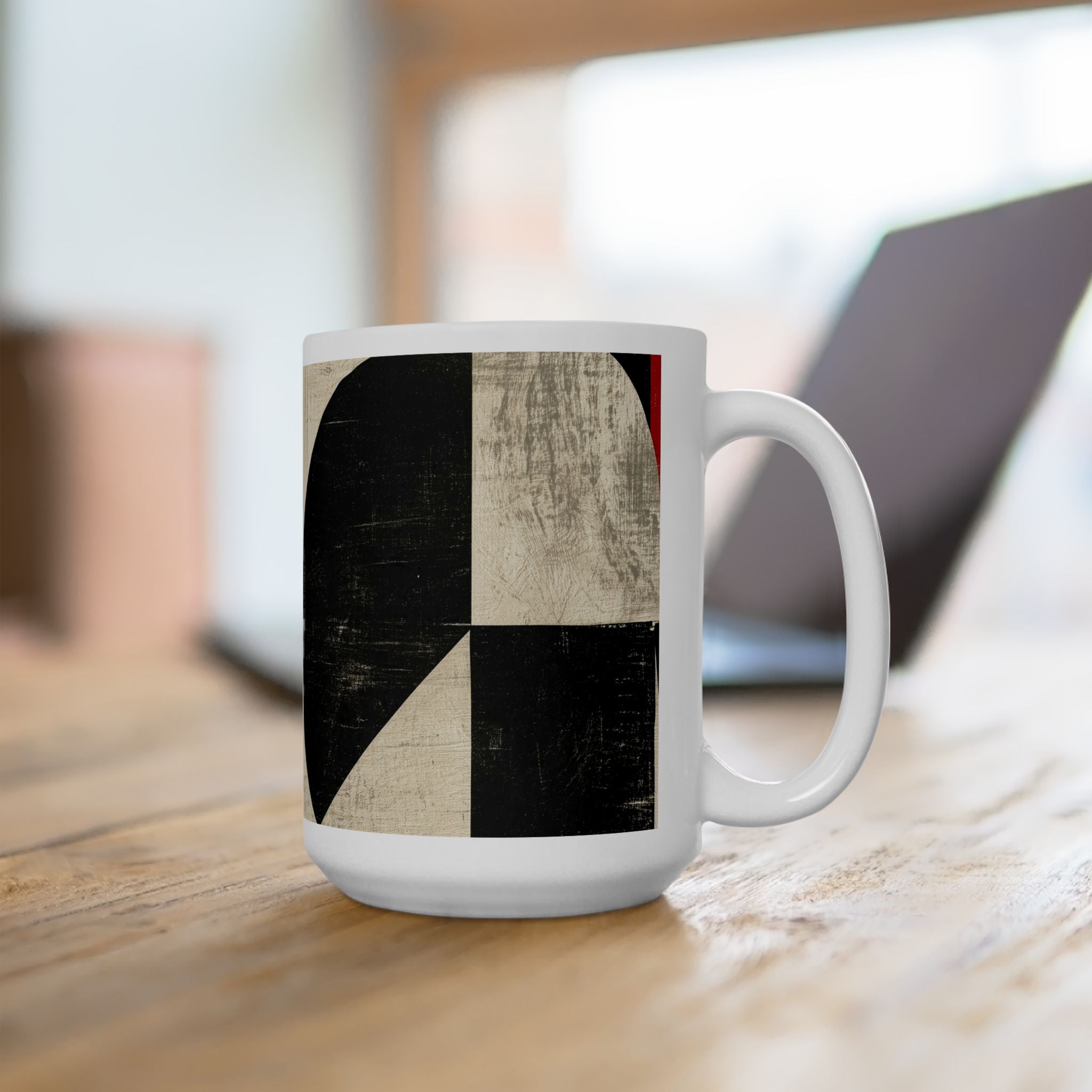 Right handle view of an 15 oz  Afrocentric geometric coffee mug featuring bold black, cream and red abstract shapes, arches, stripes and checkerboard patterns, displayed on a light wood desk with a computer laptop in the background.