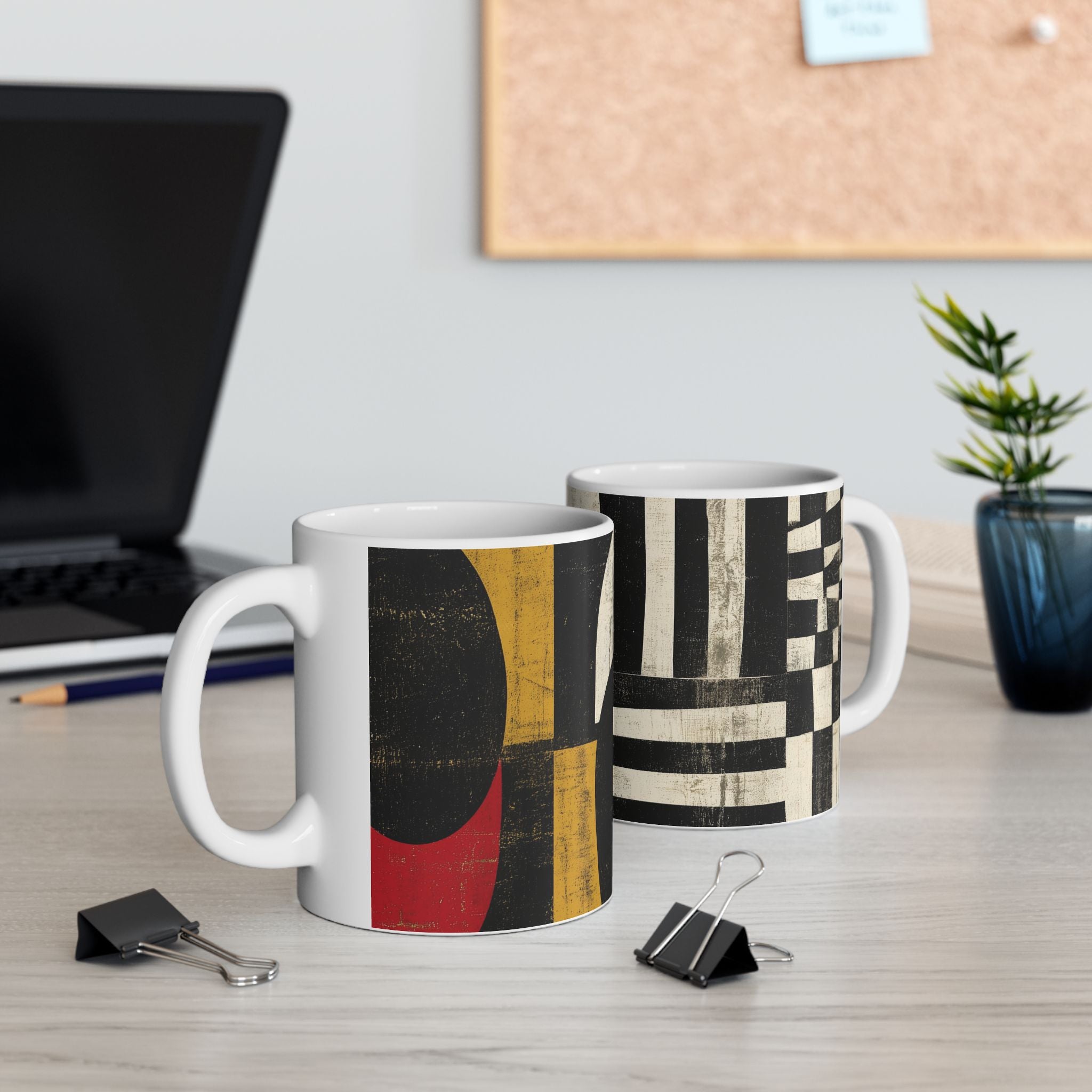 Two white ceramic mugs with abstract geometric patterns, displayed on a light wood desk with office props like a laptop, binder clips, and a small plant.