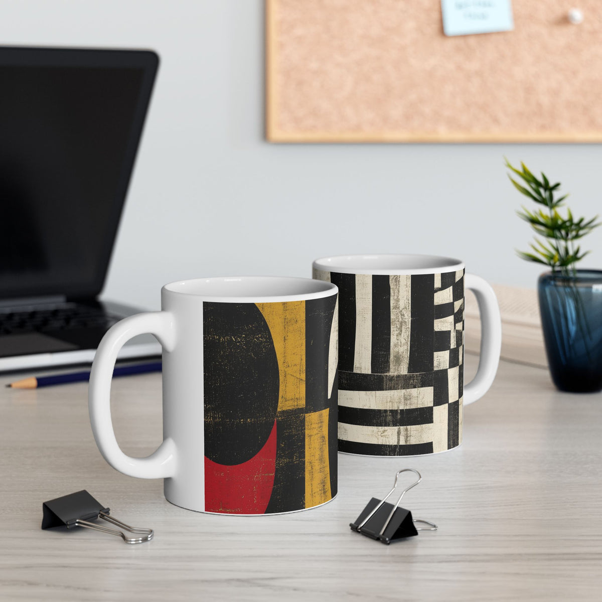 Two white ceramic mugs with abstract geometric patterns, displayed on a light wood desk with office props like a laptop, binder clips, and a small plant.