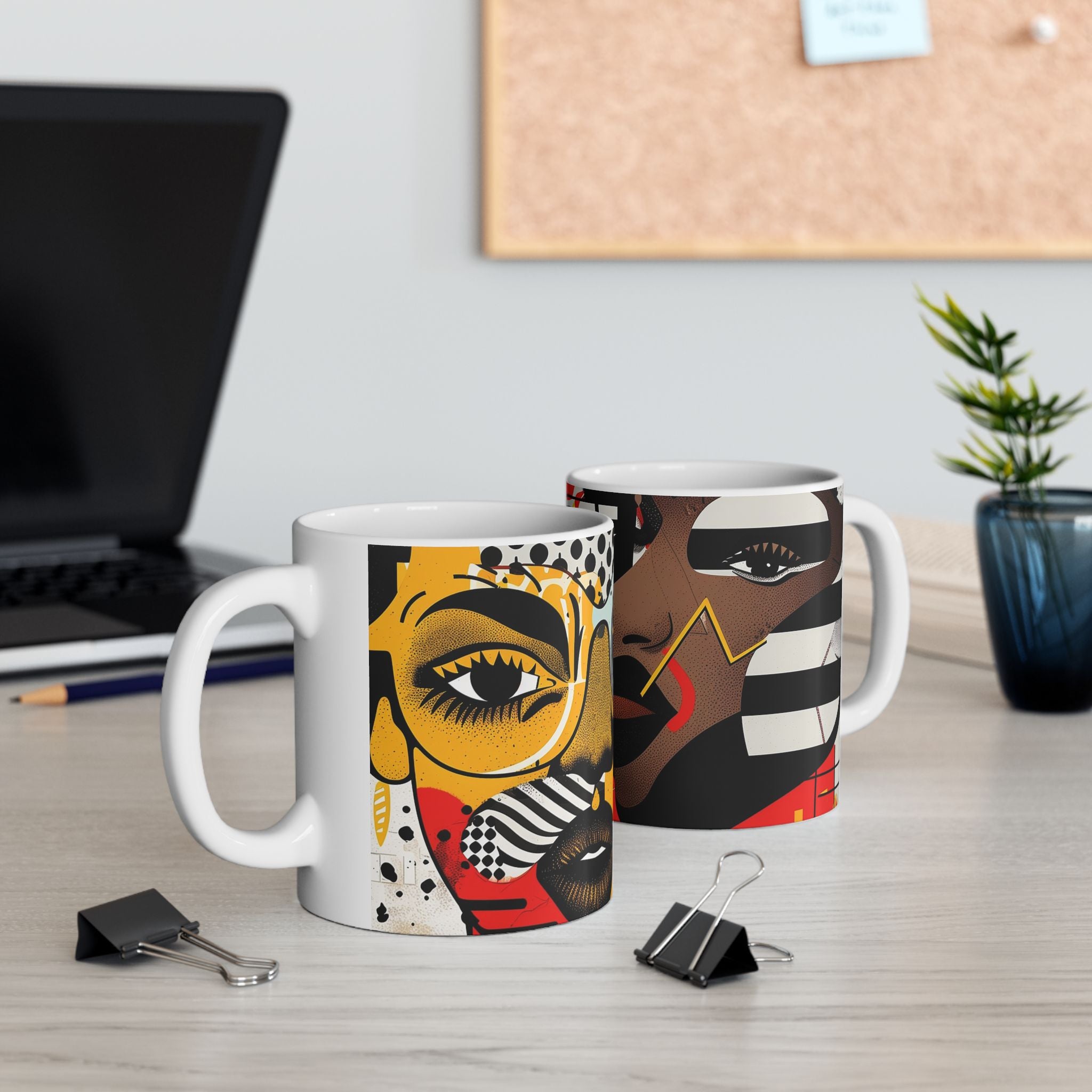 Two Soul Sisters white ceramic mugs with colorful abstract face art printed on them, displayed on a light wood desk in an office setting with a laptop, binder clips, and a small plant in the background.