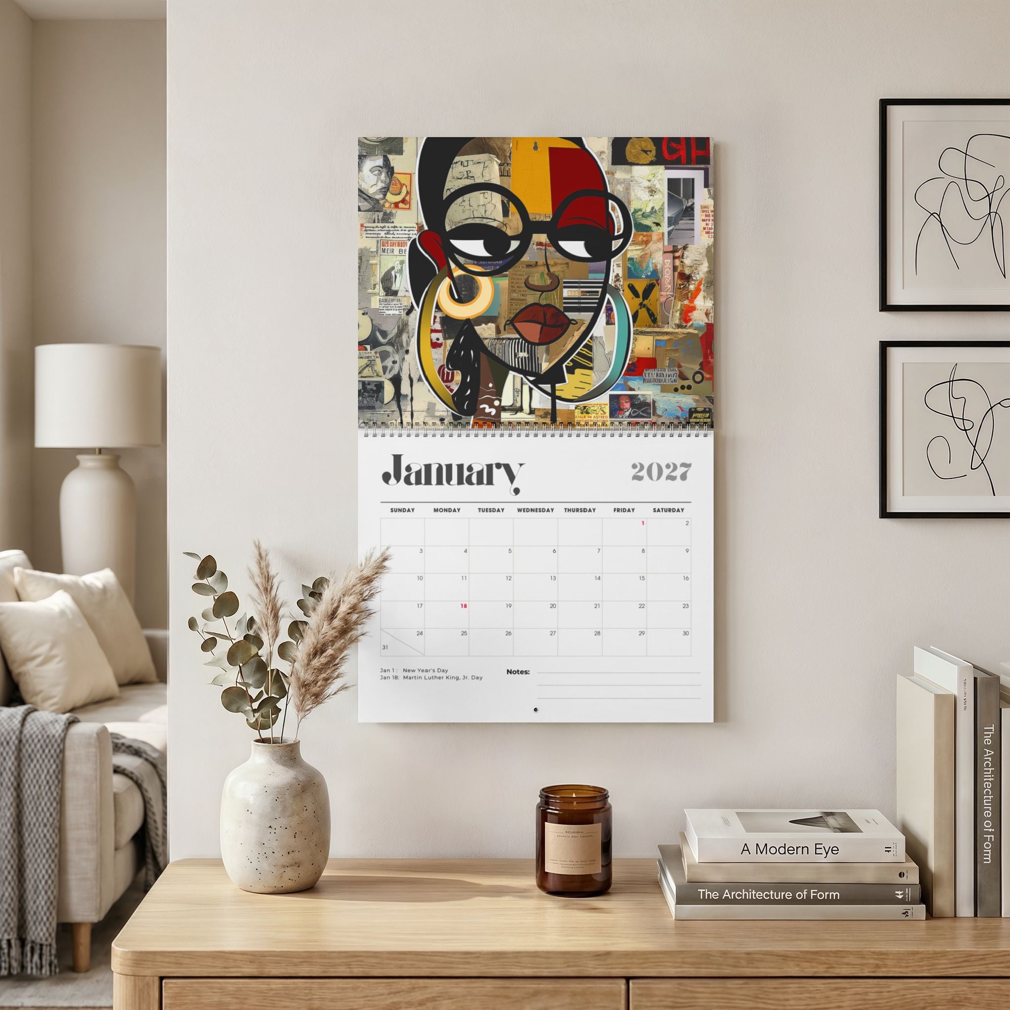 A wall-mounted 2027 January calendar featuring a colorful collage portrait of a Black female with glasses, displayed in a styled living room above a wooden console table with books, a candle, and a vase. Size 14" x "11.5"