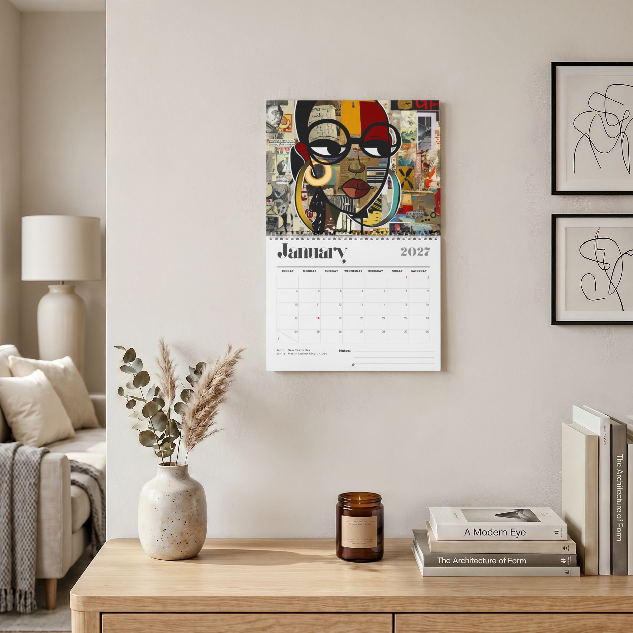 A wall-mounted 2027 January calendar featuring a colorful collage portrait of a Black female with glasses, displayed in a styled living room above a wooden console table with books, a candle, and a vase. Size 11" x 8.5"