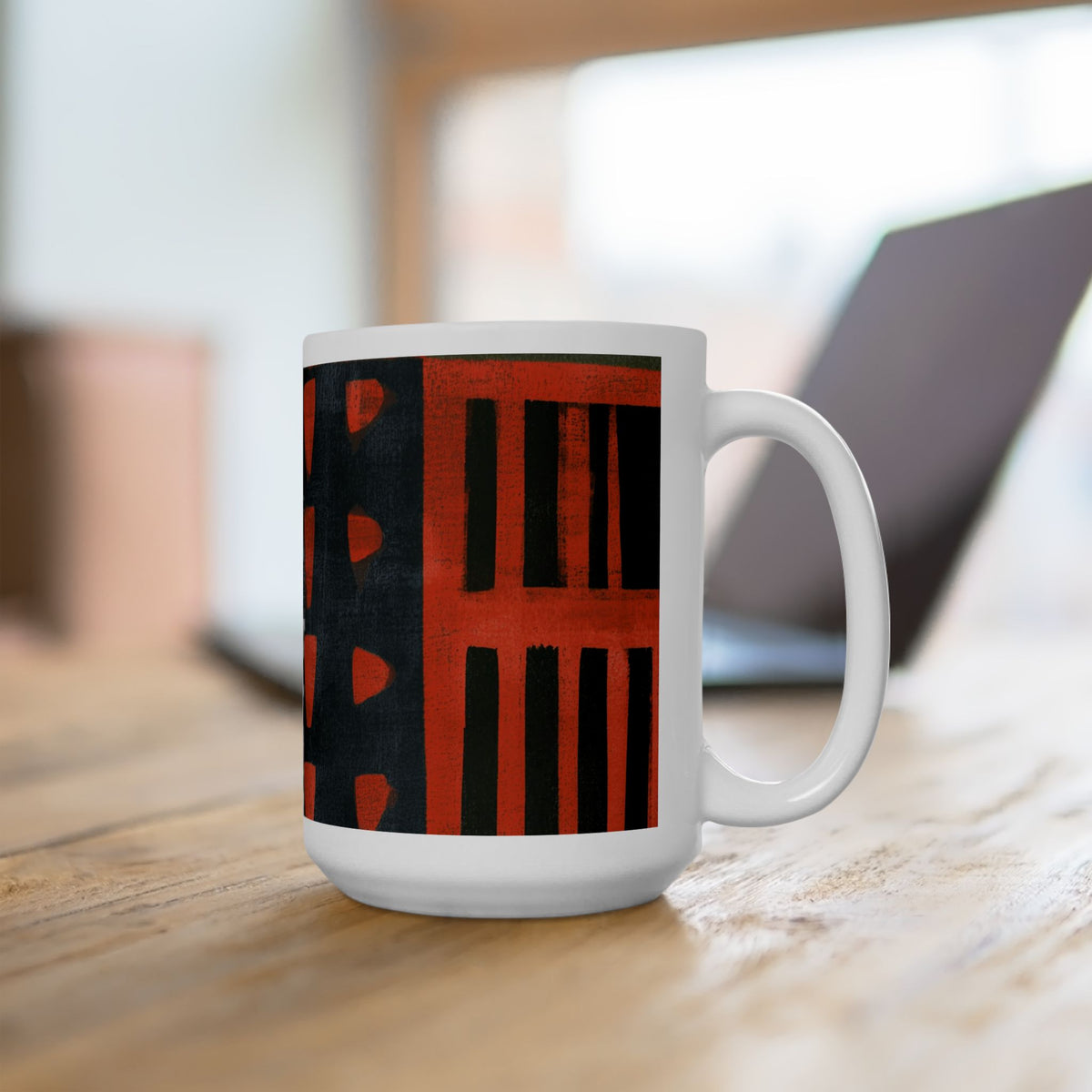 Right handle view of 15 oz. red, black, gold and cream abstract art coffee mug featuring bold geometric circles, stripes, organic shapes and a Bauhaus-inspired modern design, displayed on top of an office desk with a laptop computer in the background.