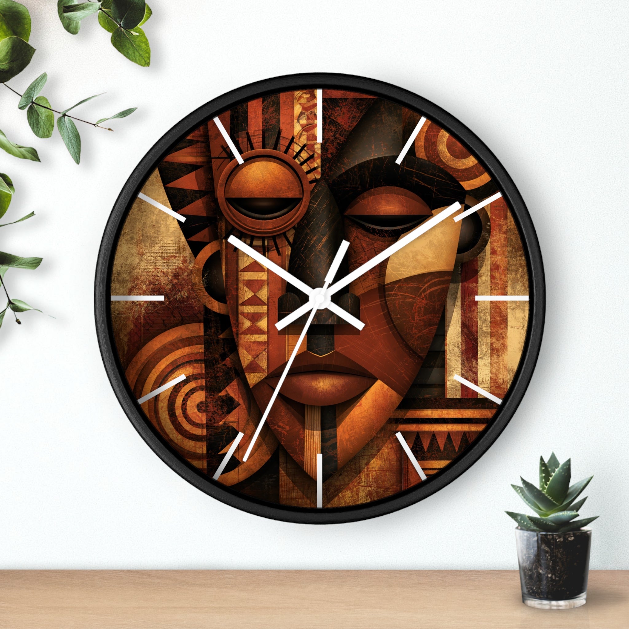 Black round wall clock with white time markings and white clock hands, featuring an abstract African-inspired mask face with geometric shapes, circular patterns, and textured details in terracotta, burnt orange, bronze, black, and warm gold tones, displayed against a white background with plant accessories.