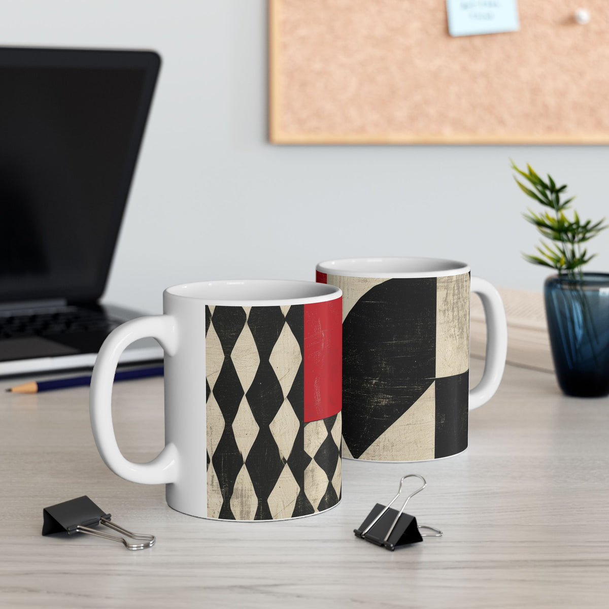 Left and right handle views of an 11 oz  Afrocentric geometric coffee mug featuring bold black, cream and red abstract shapes, arches, stripes and checkerboard patterns, displayed on a light wood desk with a computer laptop in the background.