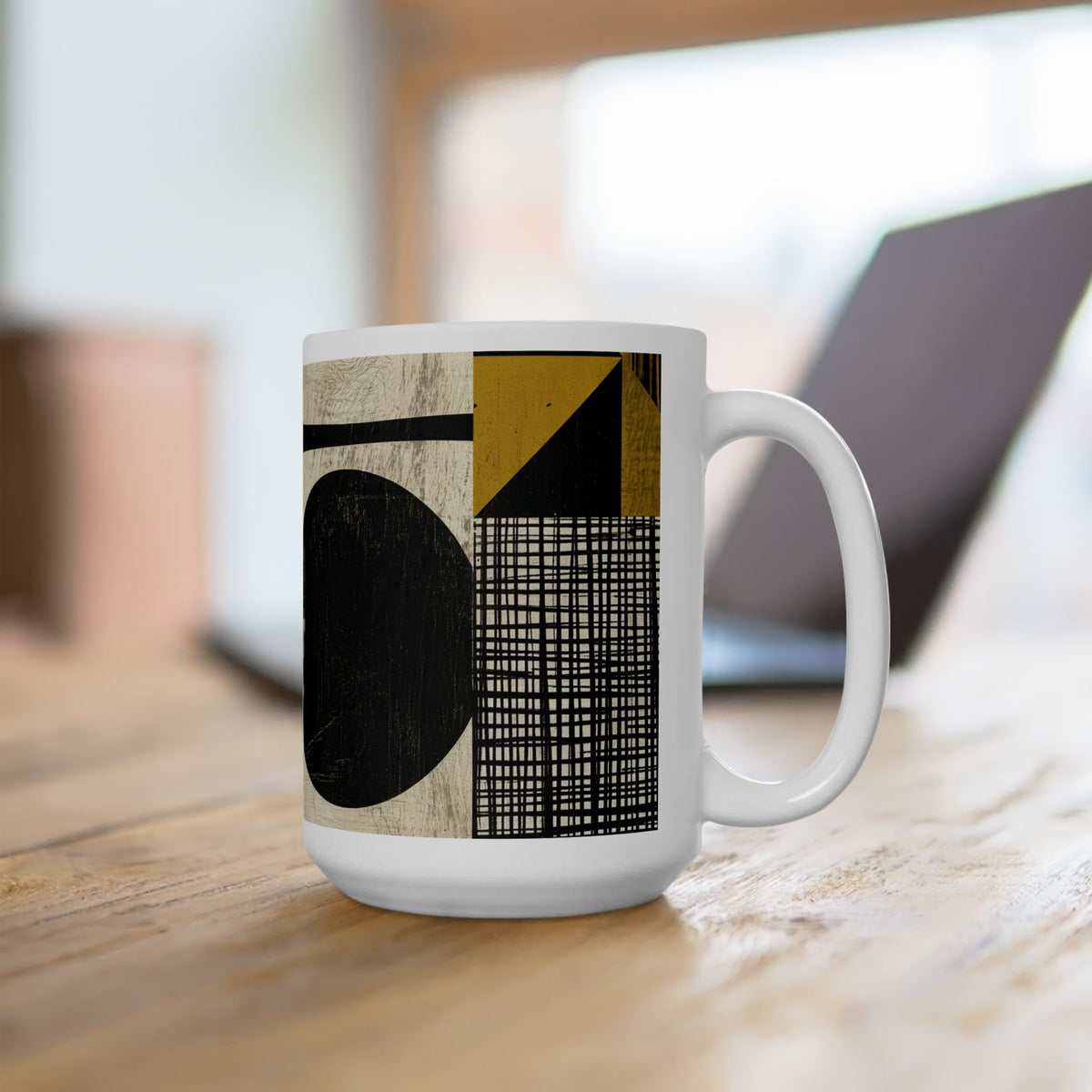 A 15 oz white ceramic mug with a modern geometric print featuring black, beige, and mustard yellow shapes, sitting on a wooden table with a blurred laptop in the background.