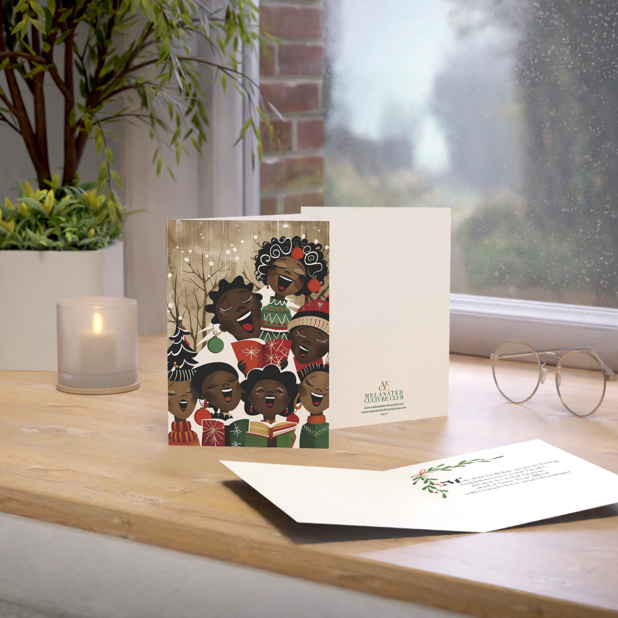 A printed greeting card and matching card are displayed on a wooden windowsill with a candle, plant, and eyeglasses nearby. The front card shows a group of illustrated Black children singing in holiday clothing, and the back card has the brand name printed near the lower right.