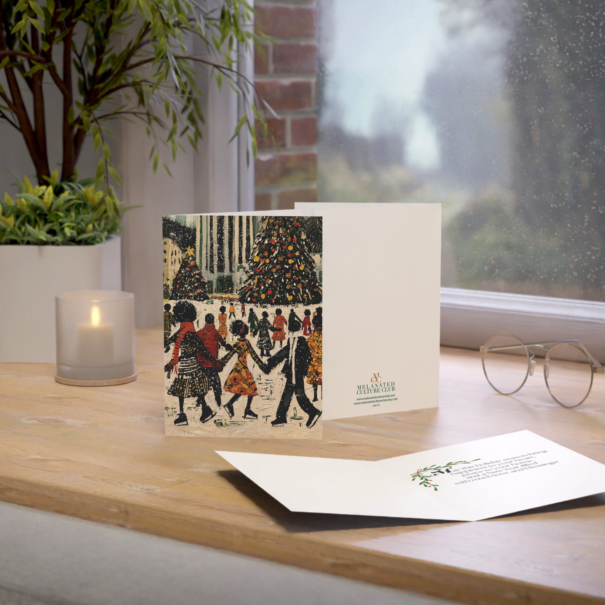 A Christmas greeting card and matching envelope displayed on a wooden windowsill, with a candle, plant, and eyeglasses nearby. The front of the card shows an illustrated winter scene with Black people skating, Christmas trees, and the back shows a small green brand logo.