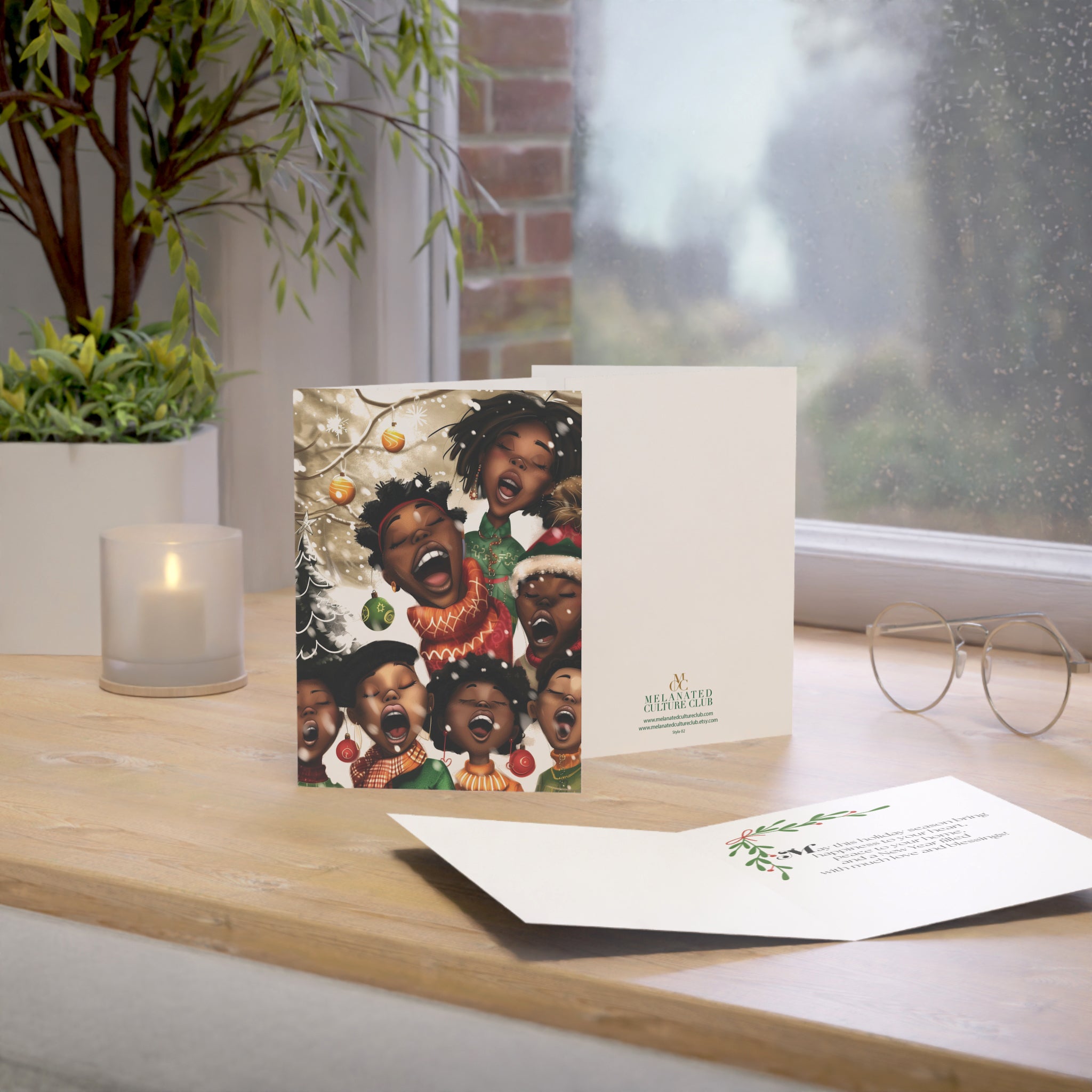 A printed greeting card and matching card are displayed on a wooden windowsill with a candle, plant, and eyeglasses nearby. The front card shows a group of illustrated Black children singing in holiday clothing, and the back card has the brand name printed near the lower right.