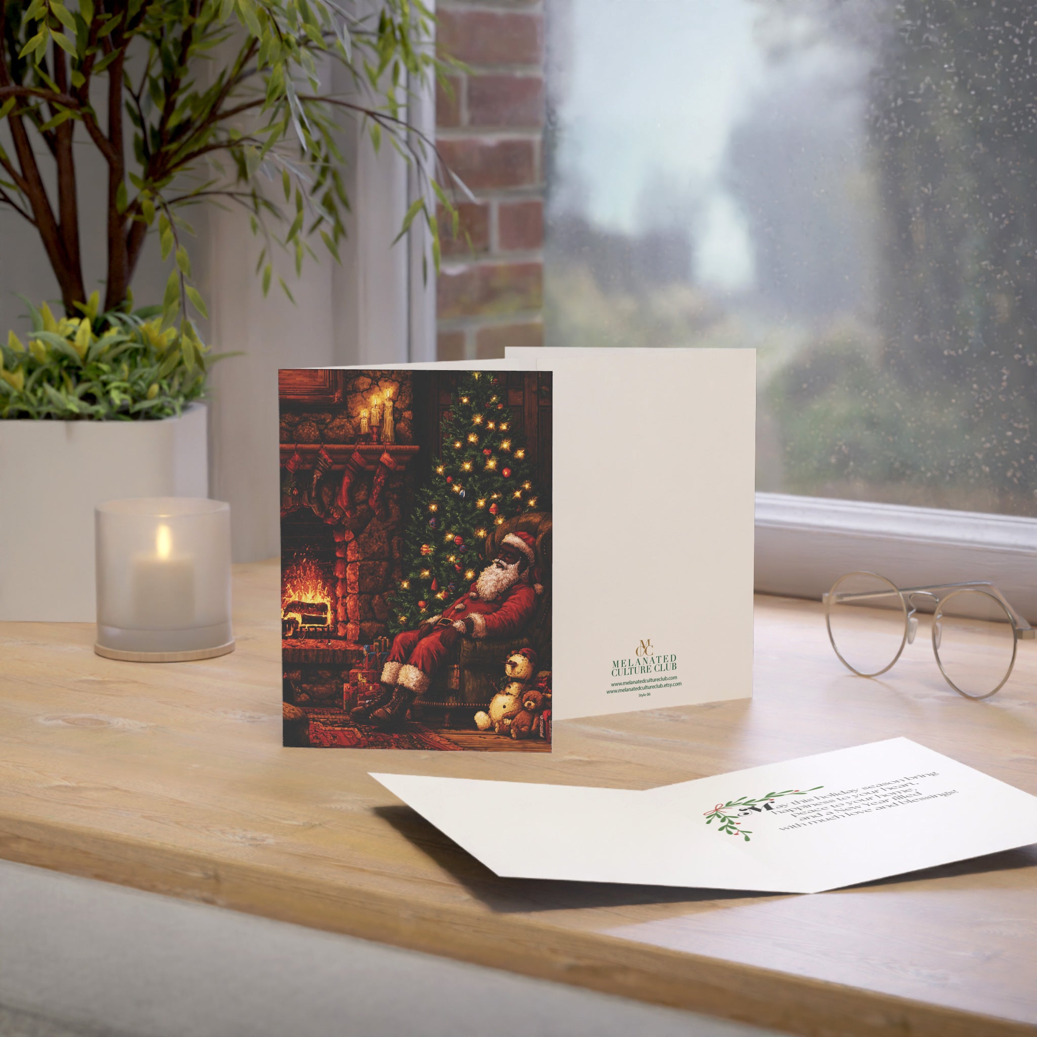 A Christmas greeting card set displayed on a wooden windowsill. The set includes a vertical card with a festive illustration of Santa Claus by a fireplace and a Christmas tree, a white envelope printed with the brand logo.