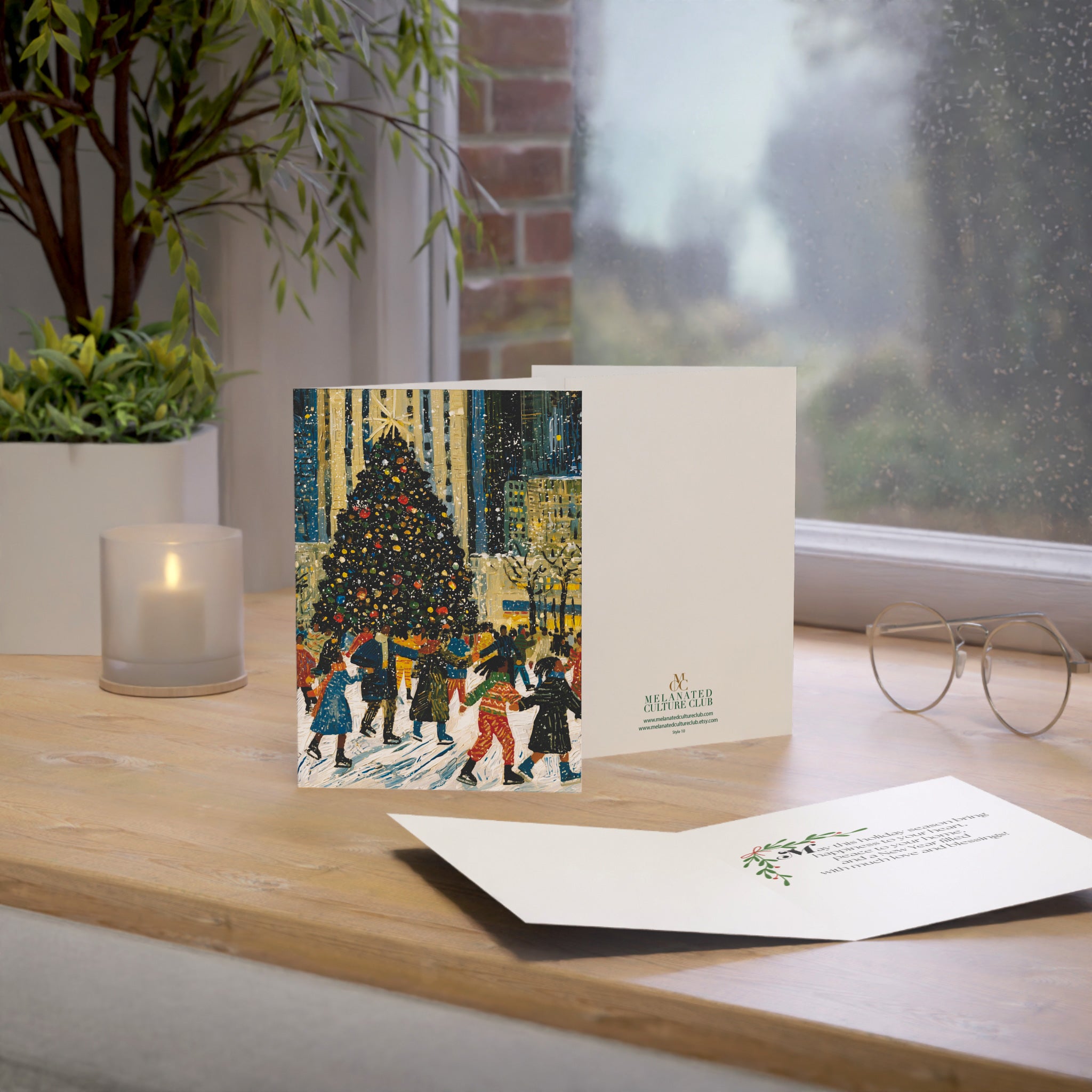 A Christmas-themed greeting card displayed on a wooden windowsill, showing a festive illustration of Black children ice skating at the Rockefeller Center with a Christmas tree. The card is shown with its white envelope and an open back insert card on a light wood surface next to a lit candle, a potted plant, and eyeglasses.