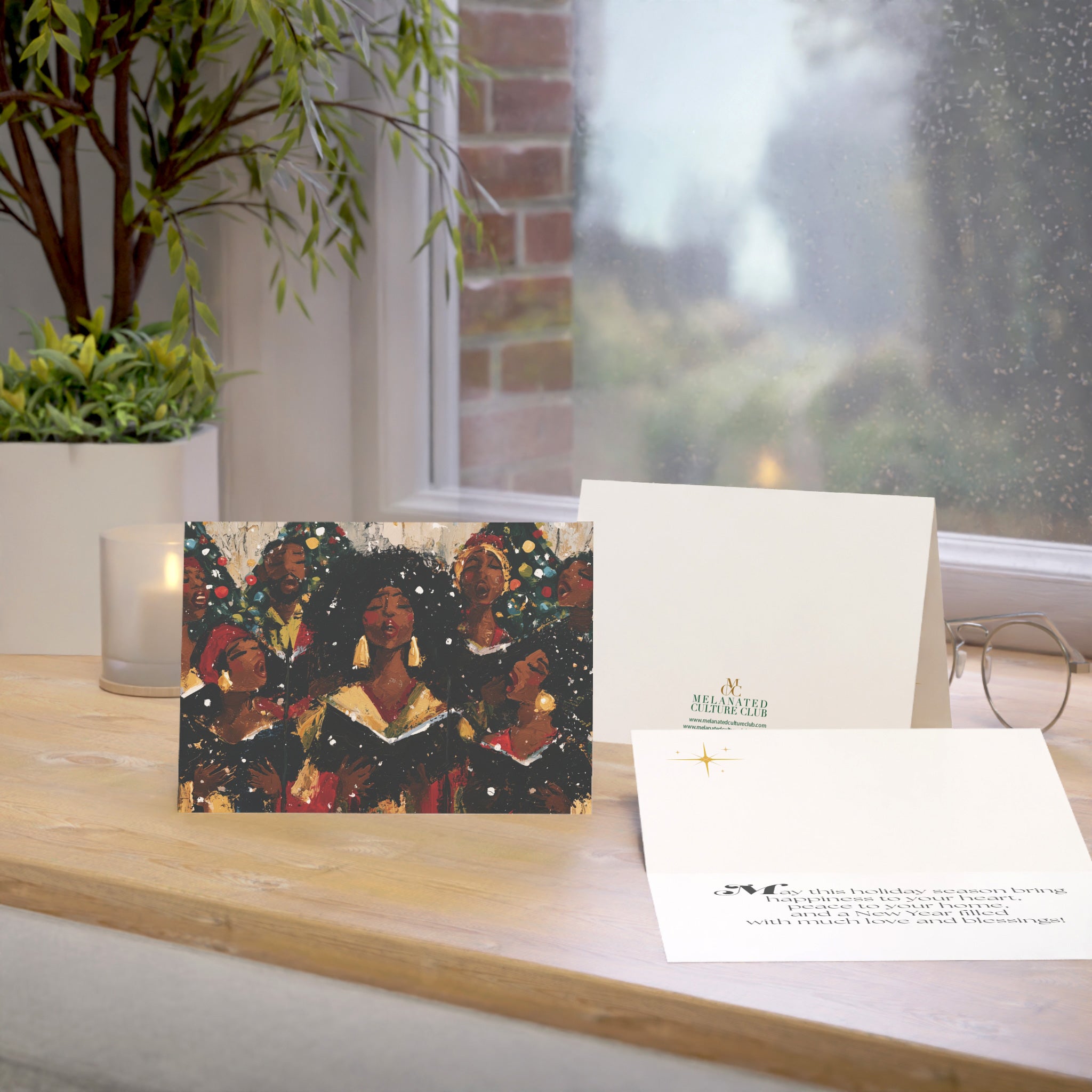 An Afrocentric holiday greeting card featuring a Black choir in black robes, displayed on a wooden windowsill beside a white envelope and a second card. A real indoor environment with a window, plant, candle, and glasses is visible in the background.