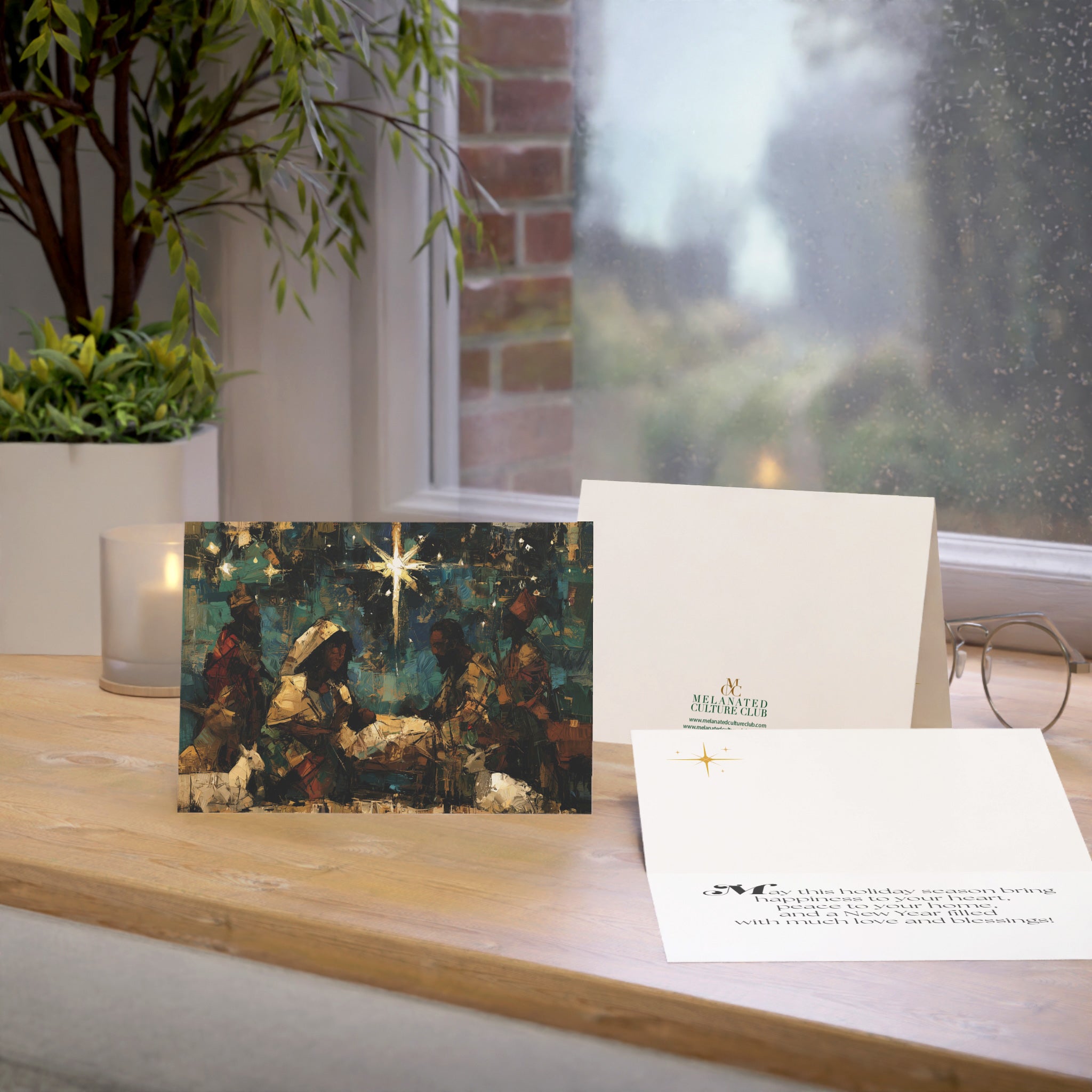 A Christmas greeting card featuring a colorful, stylized nativity scene with Black Baby Jesus, Mary, Joseph, Wise men and barn animals with a bright star in the night sky, on top of a wood surface.