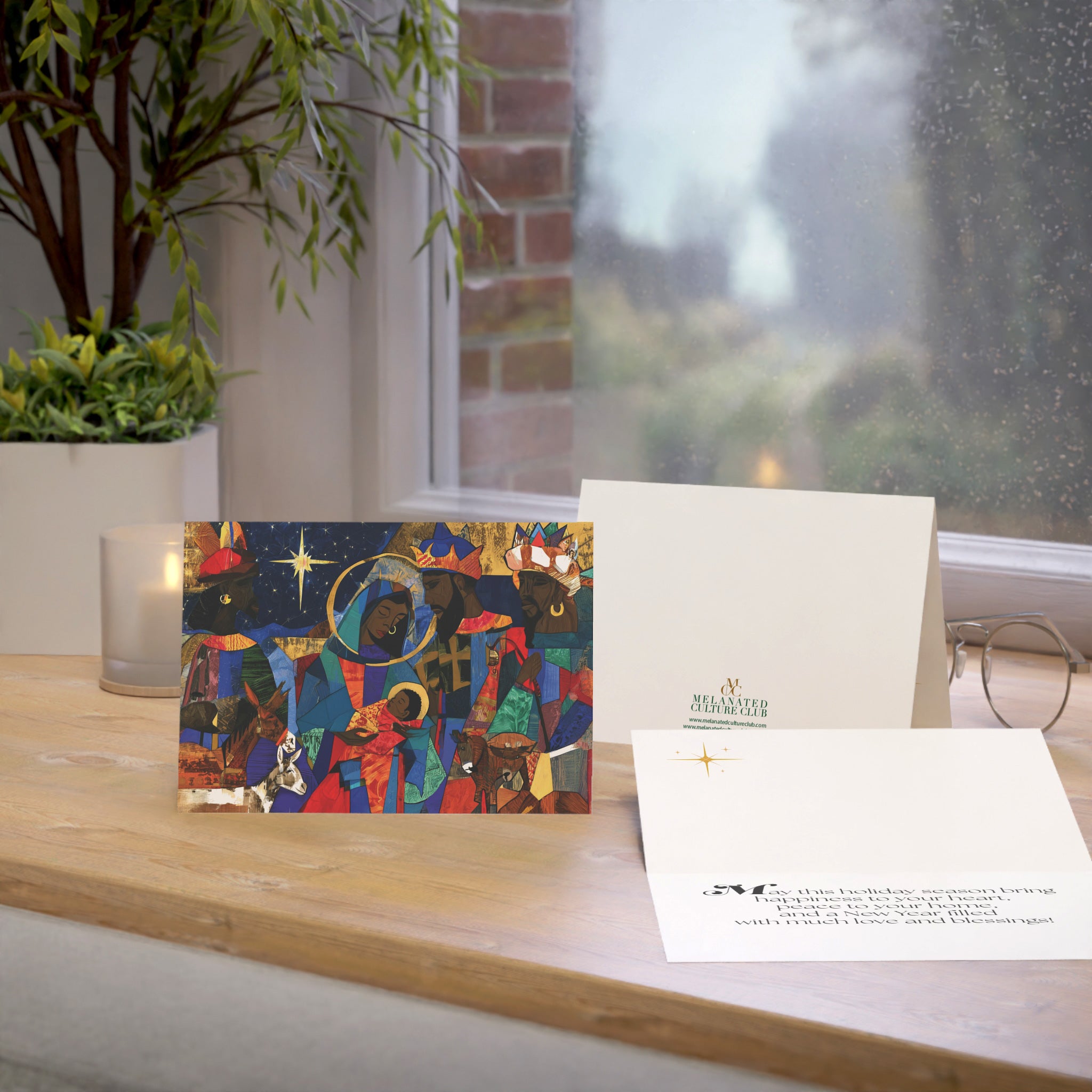 A Christmas greeting card featuring a colorful, stylized nativity scene with Black Jesus, Mary, Joseph, Wise men and barn animals with a bright star in the night sky. A potted plant and a window are visible in the softly blurred background.