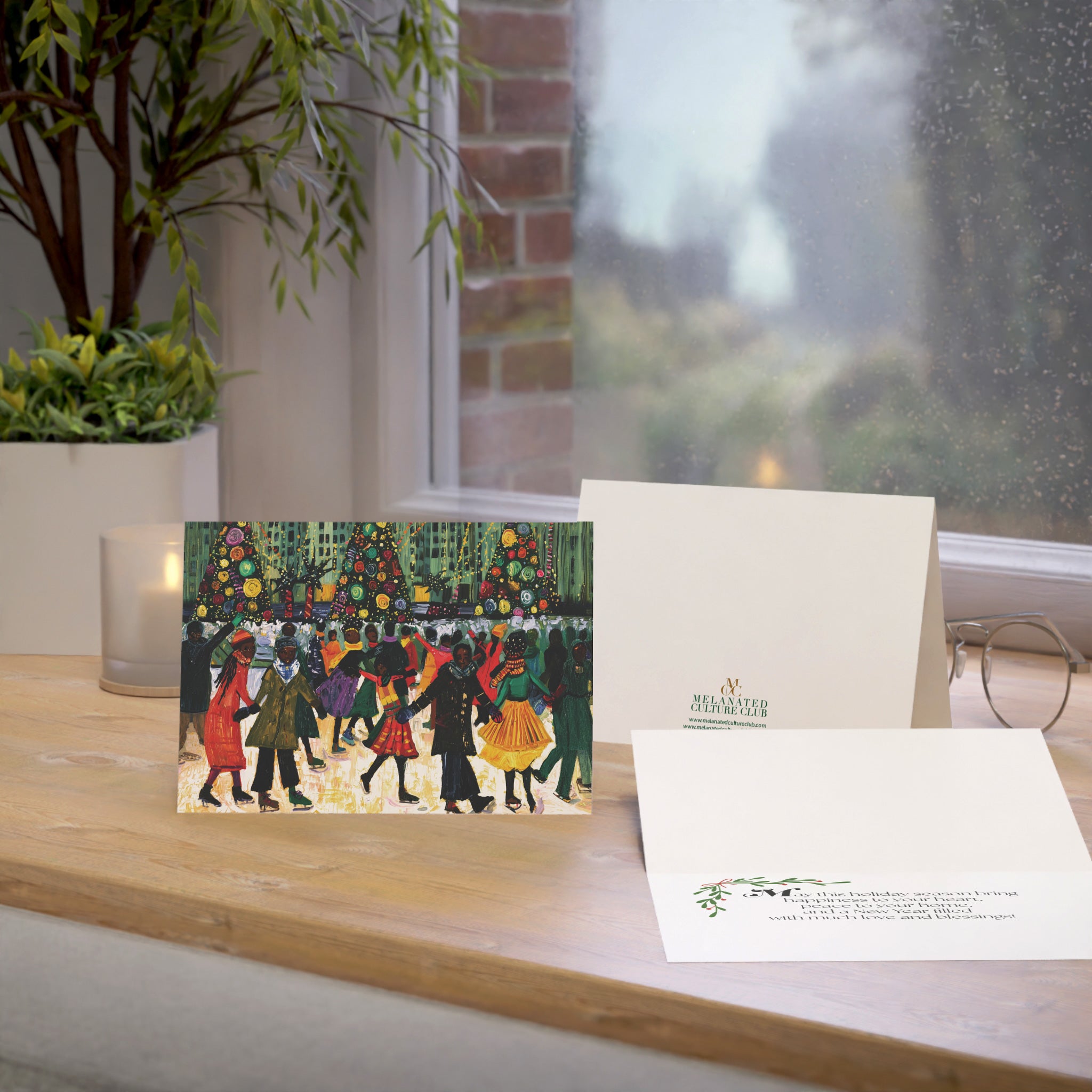 A Christmas greeting card and matching envelope displayed on a wooden windowsill, with a candle, plant, and eyeglasses nearby. The front of the card shows an illustrated winter scene with Black people skating, Christmas trees, and the back shows a small green brand logo.