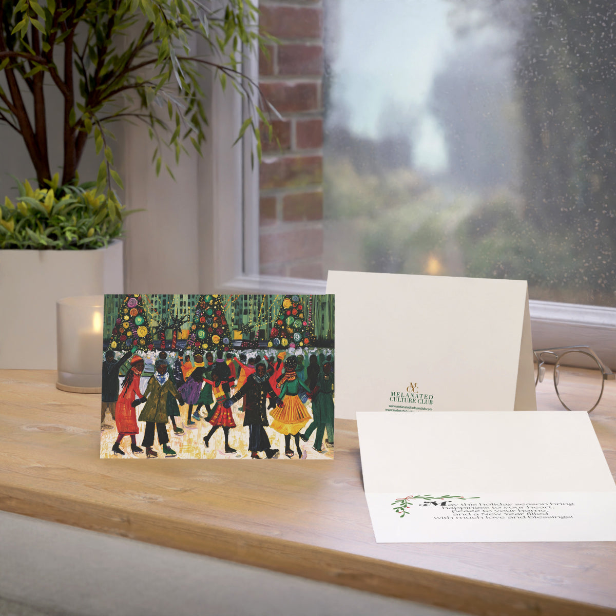 A Christmas greeting card and matching envelope displayed on a wooden windowsill, with a candle, plant, and eyeglasses nearby. The front of the card shows an illustrated winter scene with Black people skating, Christmas trees, and the back shows a small green brand logo.