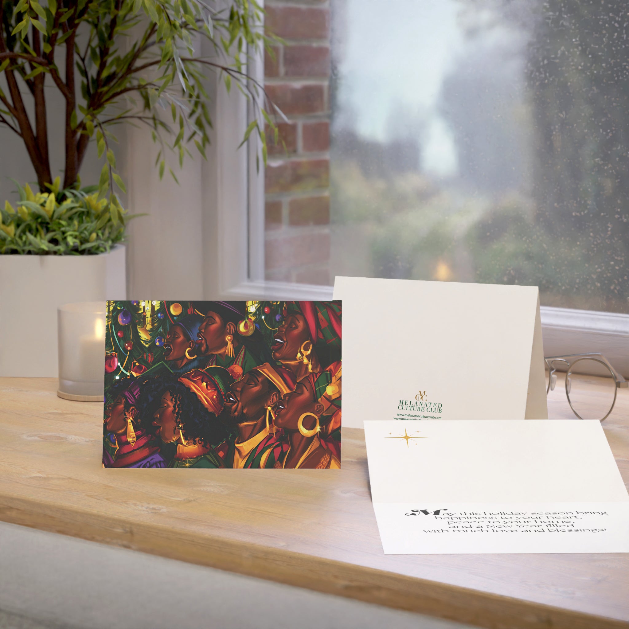 An Afrocentric holiday greeting card featuring a Black choir in black robes, displayed on a wooden windowsill beside a white envelope and a second card. A real indoor environment with a window, plant, candle, and glasses is visible in the background.