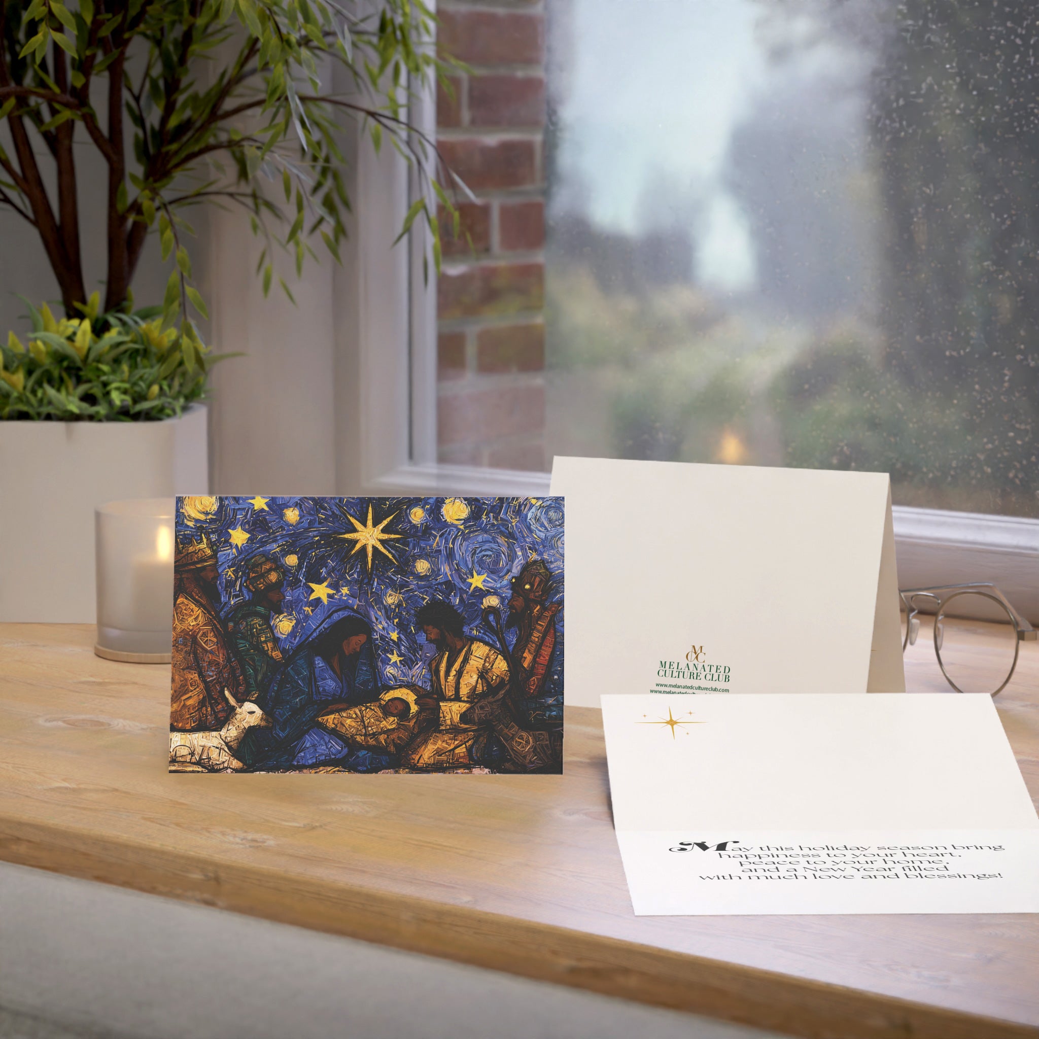 A Christmas greeting card featuring a colorful, stylized nativity scene with Black Baby Jesus, Mary, Joseph, Wise men and barn animals with a bright star in the night sky, on top of a wood surface.