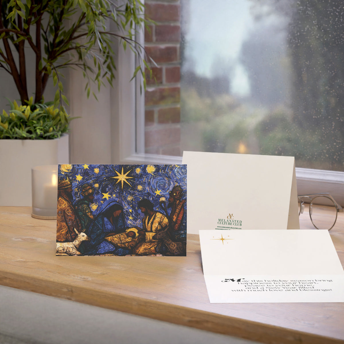 A Christmas greeting card featuring a colorful, stylized nativity scene with Black Baby Jesus, Mary, Joseph, Wise men and barn animals with a bright star in the night sky, on top of a wood surface.