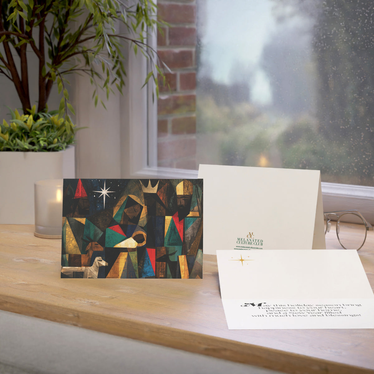 A Christmas greeting card featuring a colorful, stylized nativity scene with Black Baby Jesus, Mary, Joseph, Wise men and barn animals with a bright star in the night sky, on top of a wood surface.