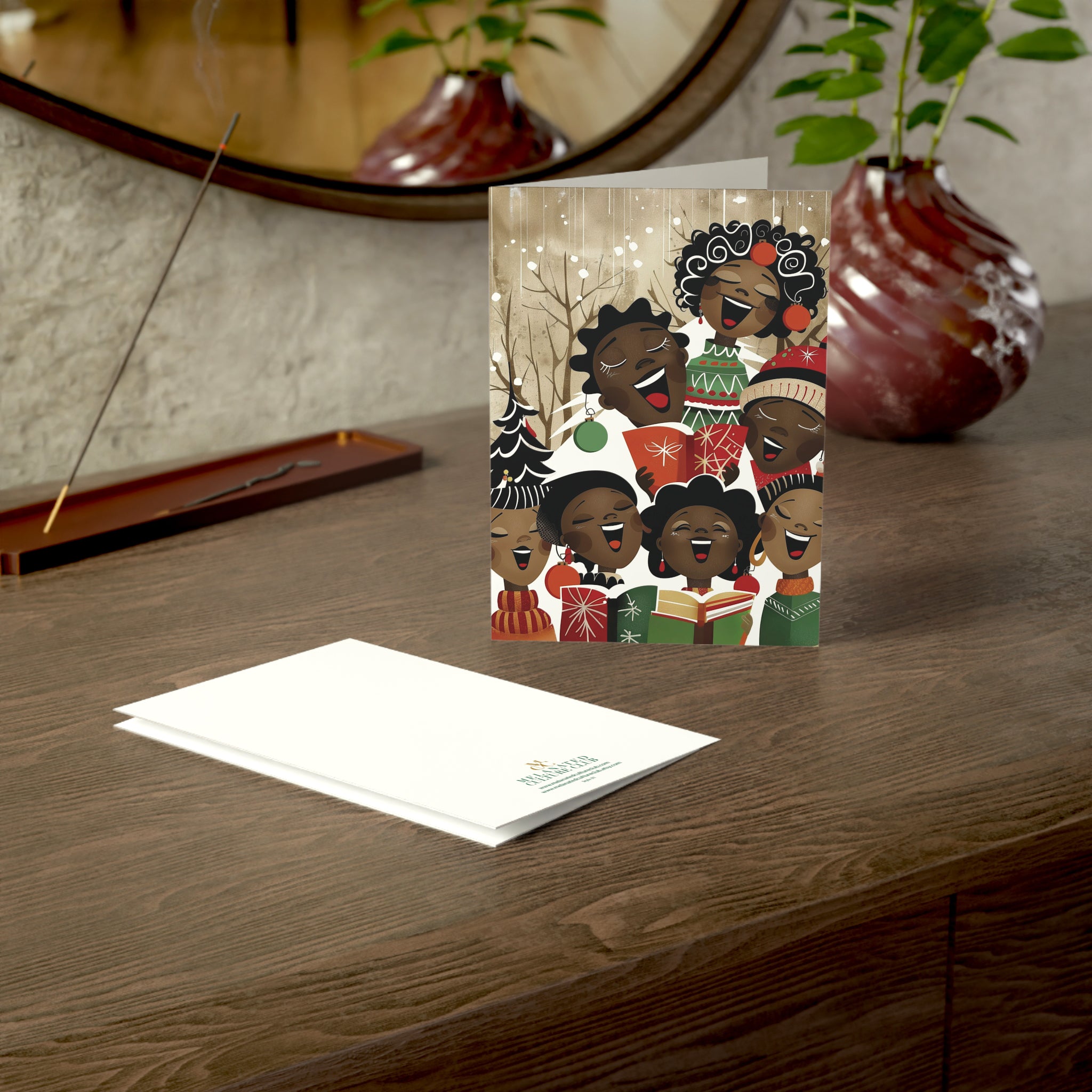 A Christmas-themed art print card featuring a group of Black children singing Christmas carols, displayed on a wooden table next to its white envelope. The background shows a warm, styled interior with a mirror, plants, and a vase.