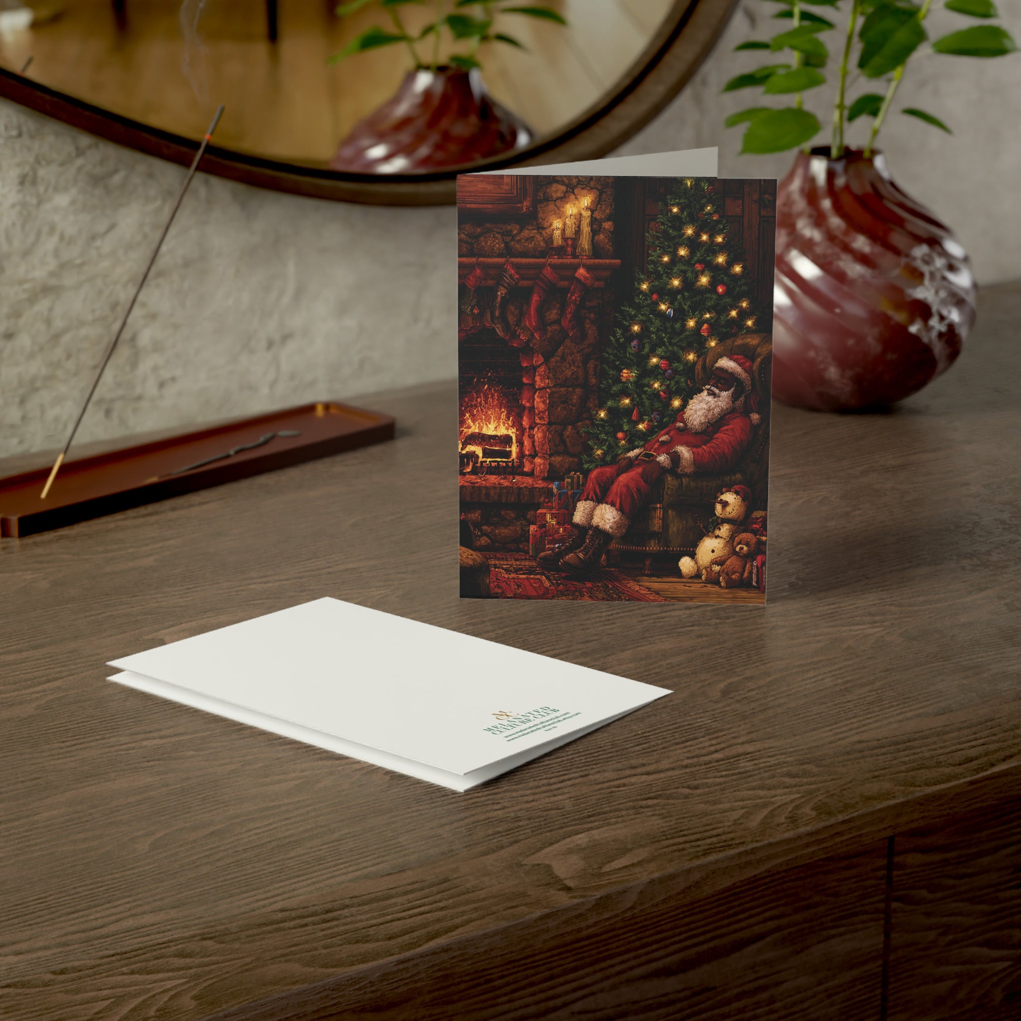 A Christmas greeting card featuring Black Santa Claus sitting by a decorated tree in a cozy living room, displayed on a wooden table next to its white envelope. The card is set in a warm, styled interior with a mirror, plants, and a vase in the background.