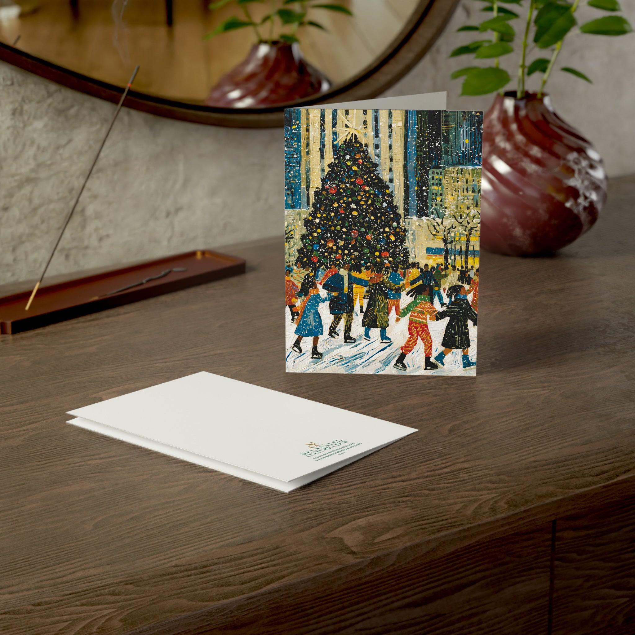 A greeting card standing upright on a wooden table, showing an illustration of Black children ice skating at the Rockefeller Center skating around a decorated Christmas tree. A white card envelope lies flat in front of it.