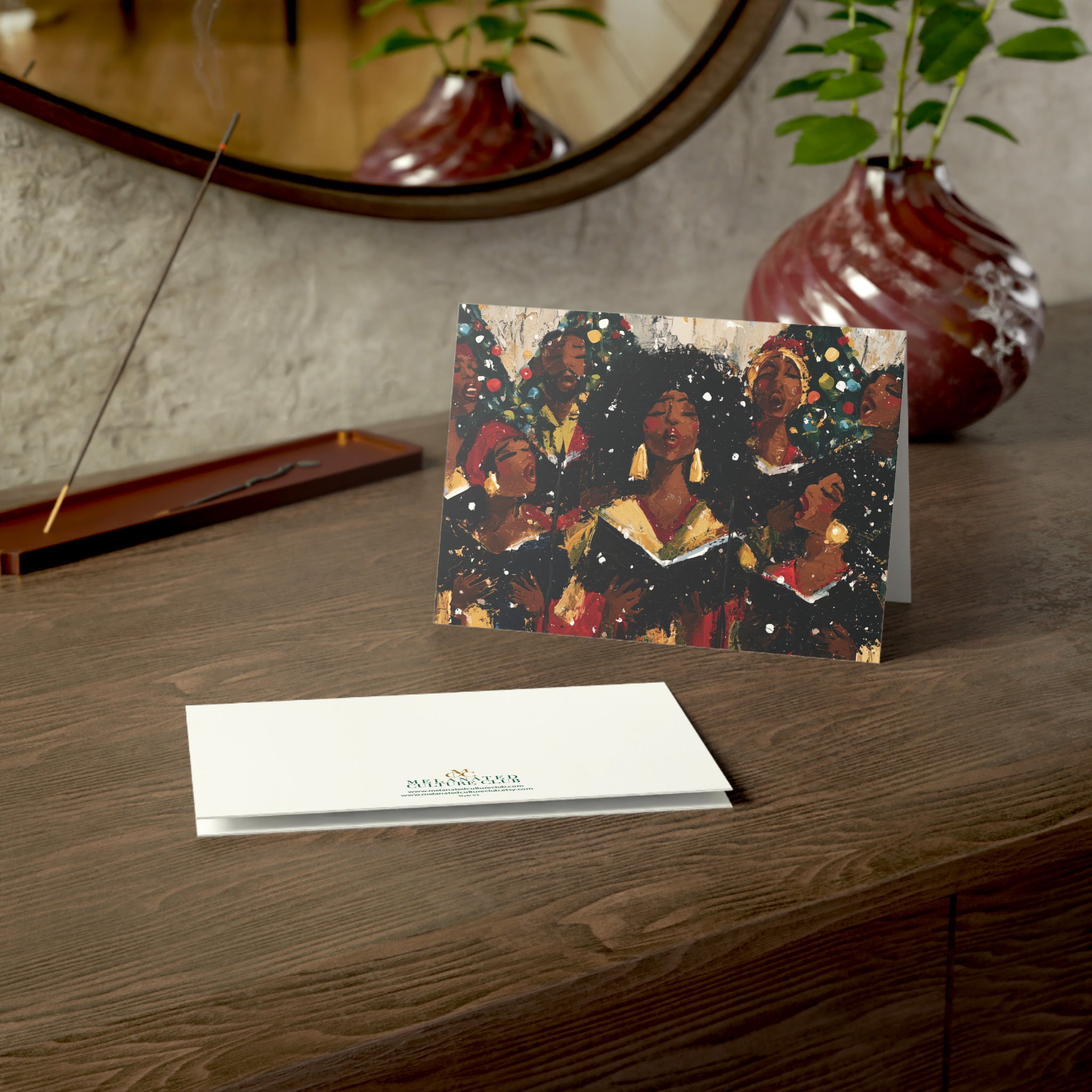 An Afrocentric holiday greeting card featuring a Black choir in black robes, , standing on a dark wooden table next to a white envelope printed with the brand logo. The background shows a textured wall, a mirror, and potted plants.