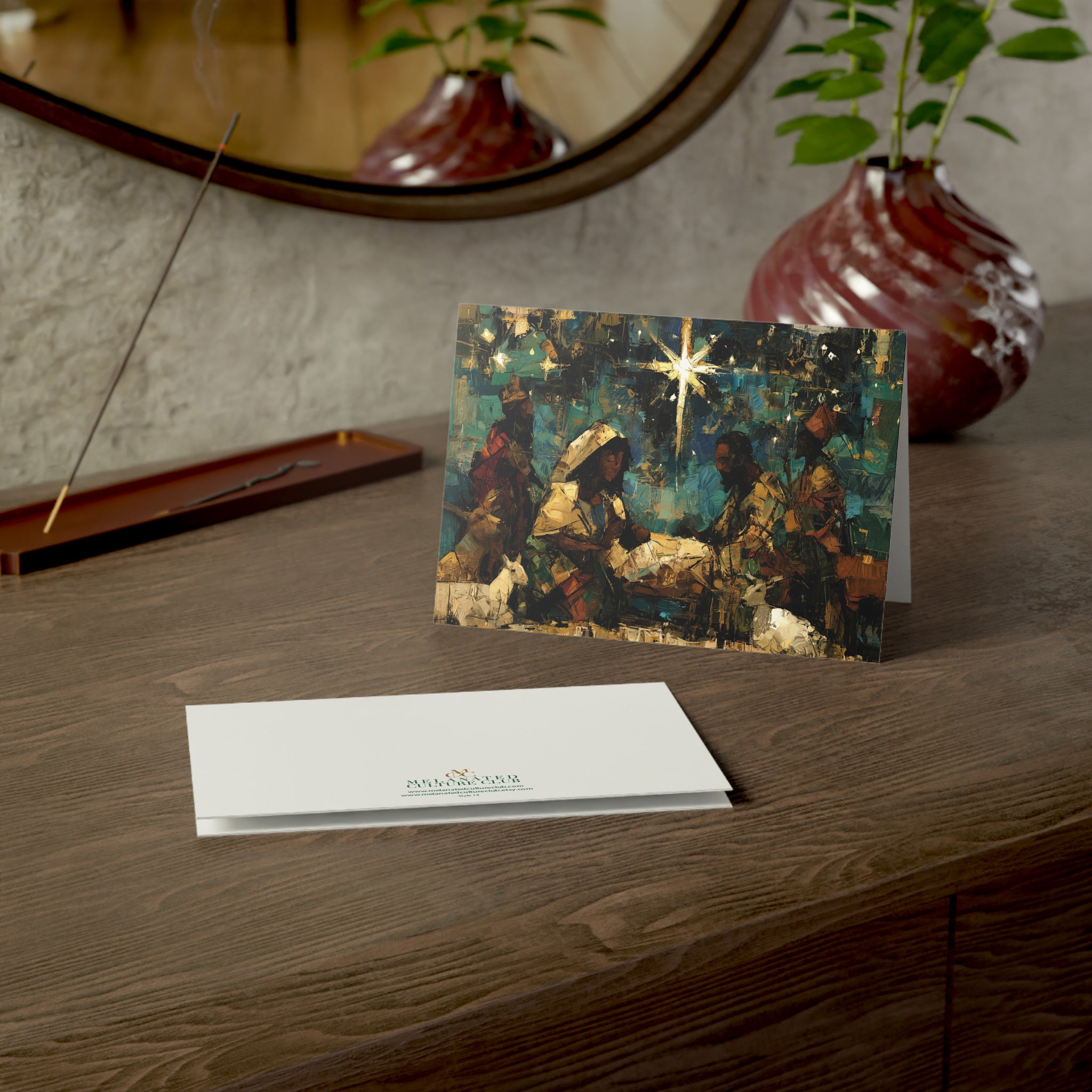 A Christmas greeting card featuring a colorful, stylized nativity scene with Black Baby Jesus, Mary, Joseph, Wise men and barn animals with a bright star in the night sky, on top of a wood surface.