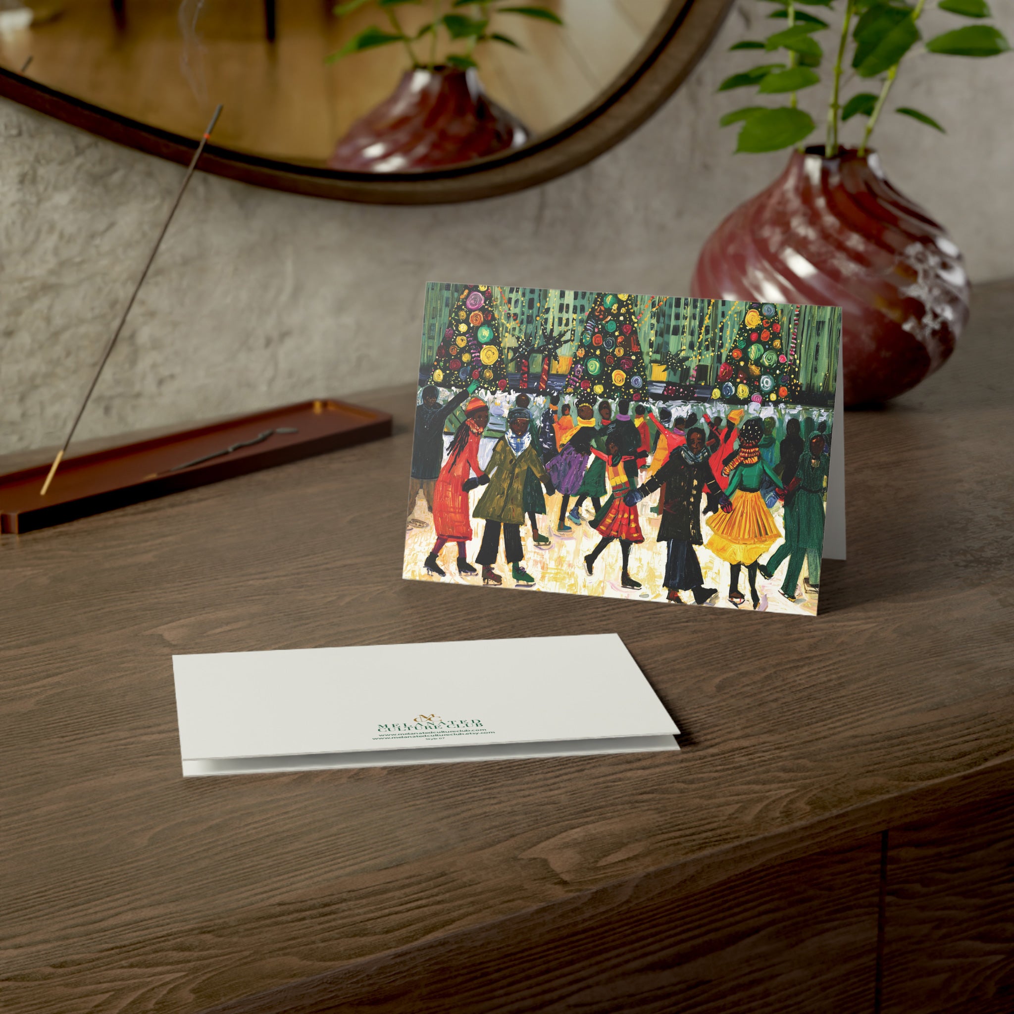 A horizontal greeting card with an abstract illustration of Black people ice skating with Christmas trees in the background at the Rockefeller Center, stands on a wooden table, next to a folded white card with a small printed brand logo.