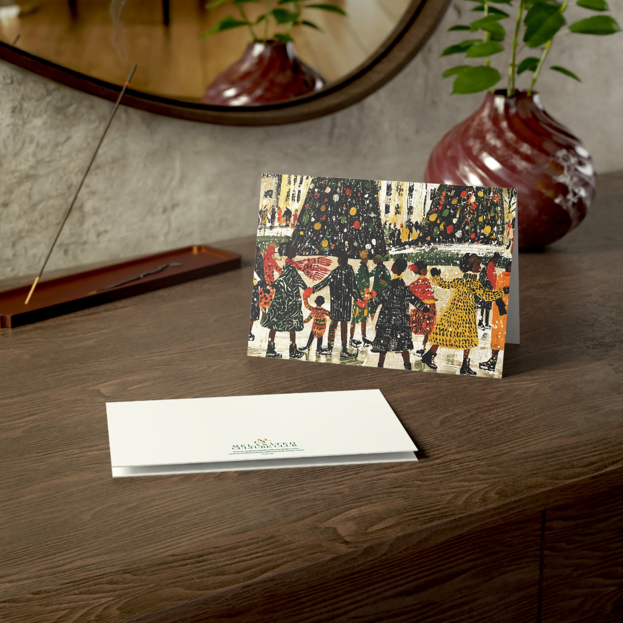 A Christmas greeting card and matching envelope displayed on a wooden windowsill, with a candle, plant, and eyeglasses nearby. The front of the card shows an illustrated winter scene with Black people skating, Christmas trees, and the back shows a small green brand logo.